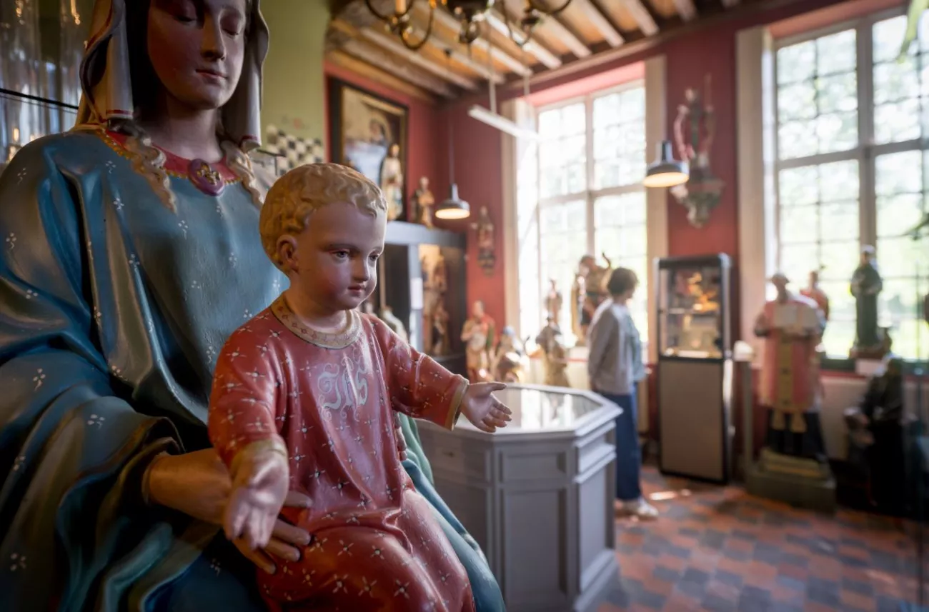 Gran estatua pintada de la Virgen María sosteniendo al Niño Jesús en primer plano, con estatuas religiosas adicionales, vitrinas de cristal y un visitante visible en el fondo del interior del museo con paredes rojas y vigas de madera en el techo.