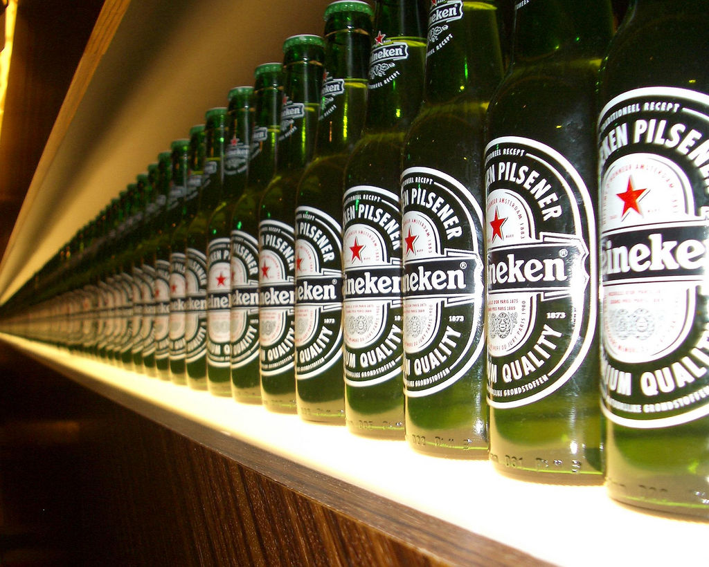 Row of Heineken green glass bottles with red star logo on a lit shelf, extending into the distance with perspective effect
