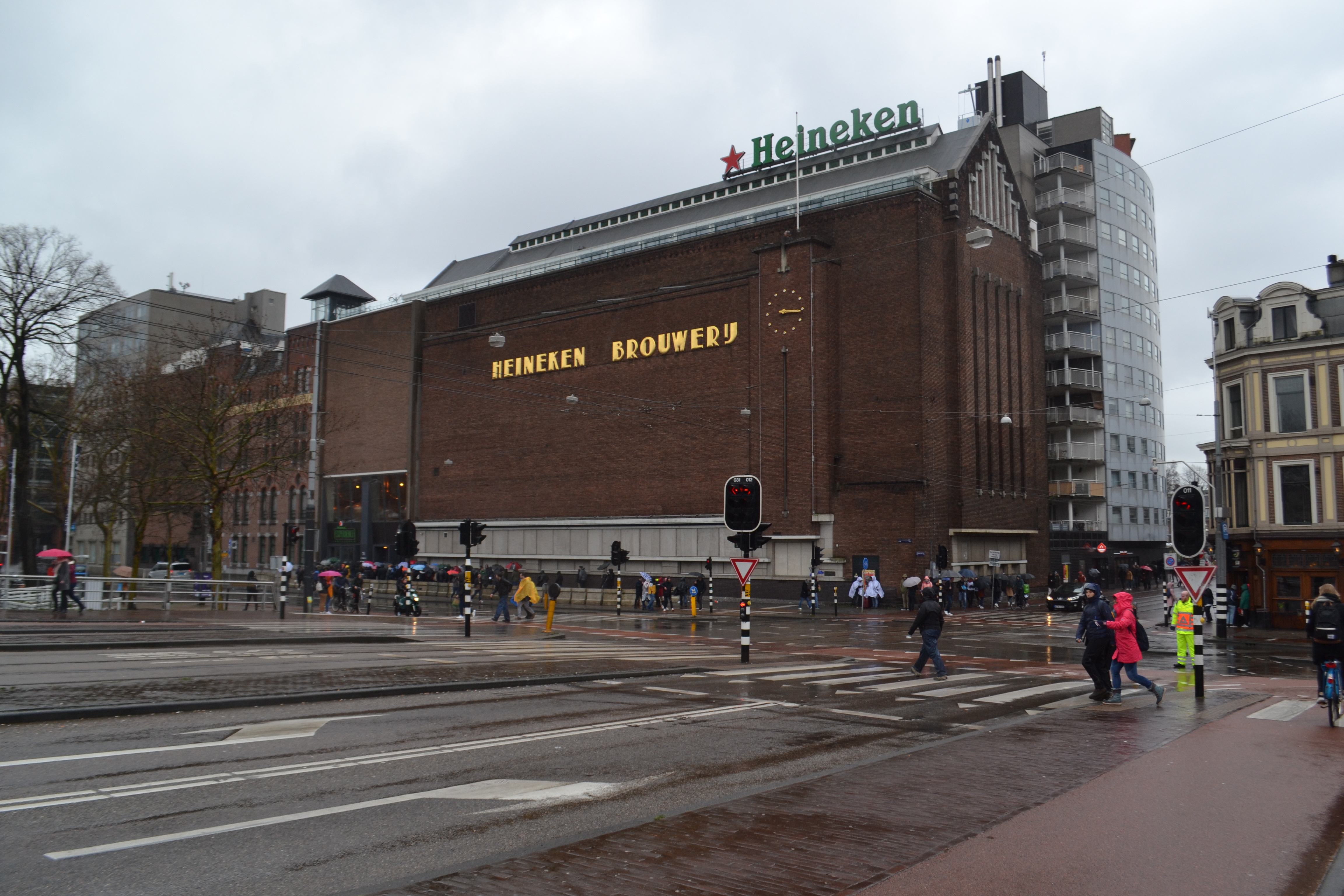 Exterior of the Heineken Experience building, a former brewery with large 'Heineken' and 'Heineken Brouwerij' signage on a brick facade, viewed from across a wet city street with pedestrians and traffic lights