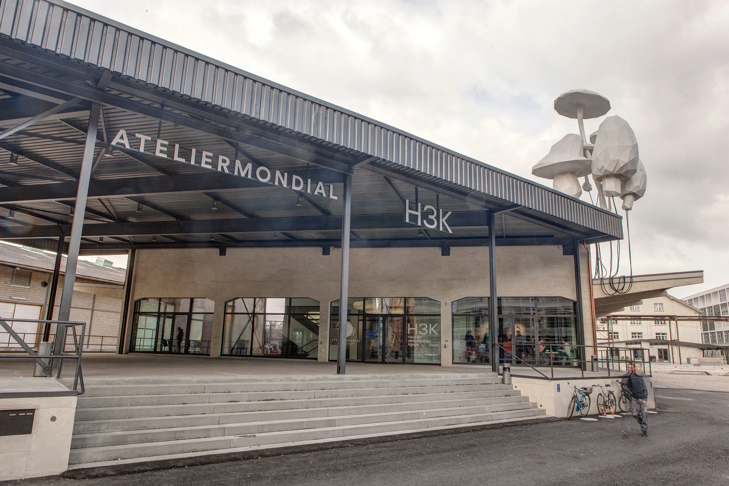 Widok z zewnątrz na fasadę nowoczesnego budynku przemysłowego z dużym napisem „ATELIER MONDIAL” i „H3K”, szklanymi oknami, szerokimi betonowymi schodami oraz wysoką instalacją rzeźbiarską po prawej stronie.