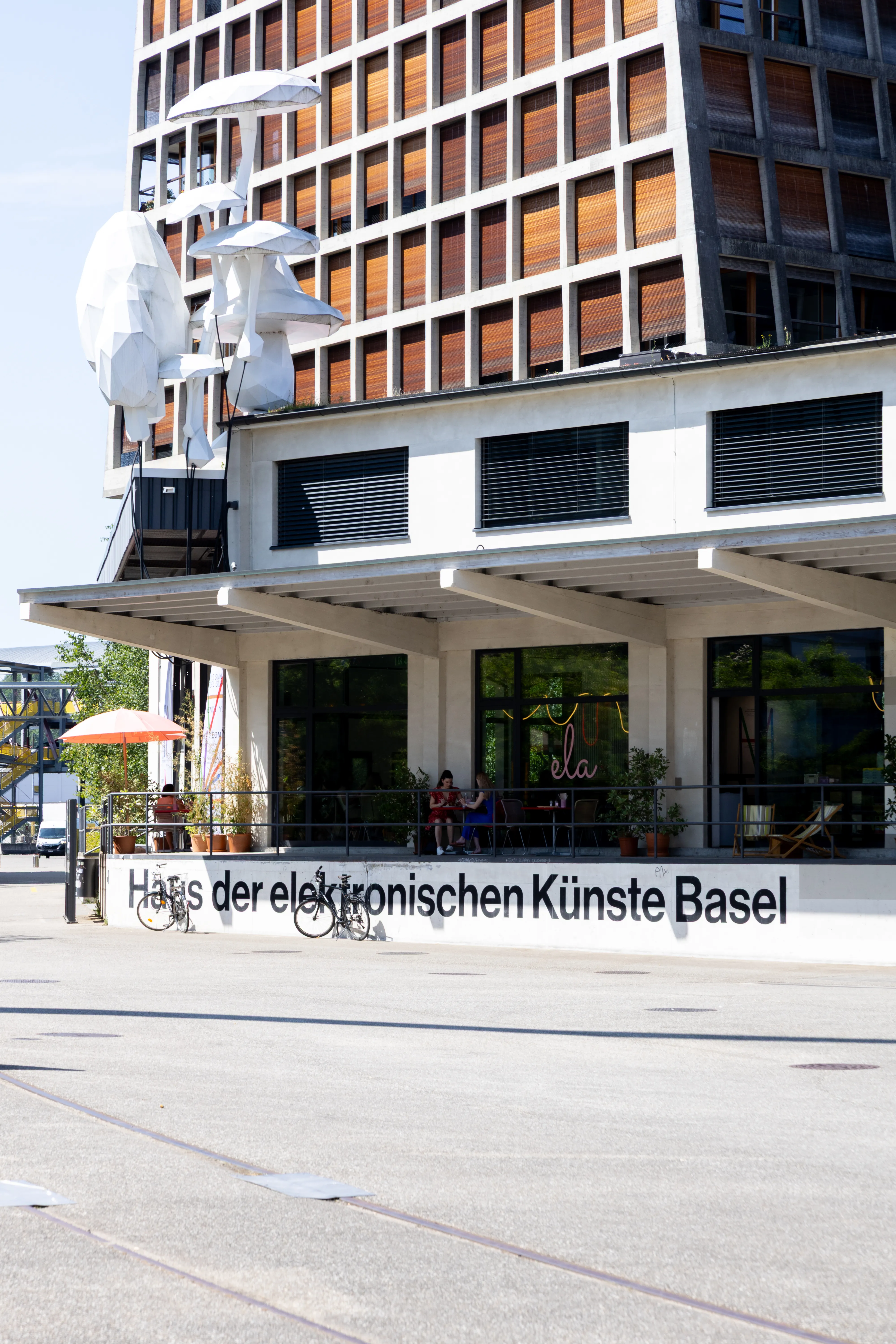 Widok z zewnątrz na budynek Haus der elektronischen Künste Basel z białą fasadą, dużymi oknami, wystającym zadaszeniem i miejscami do siedzenia na zewnątrz.
