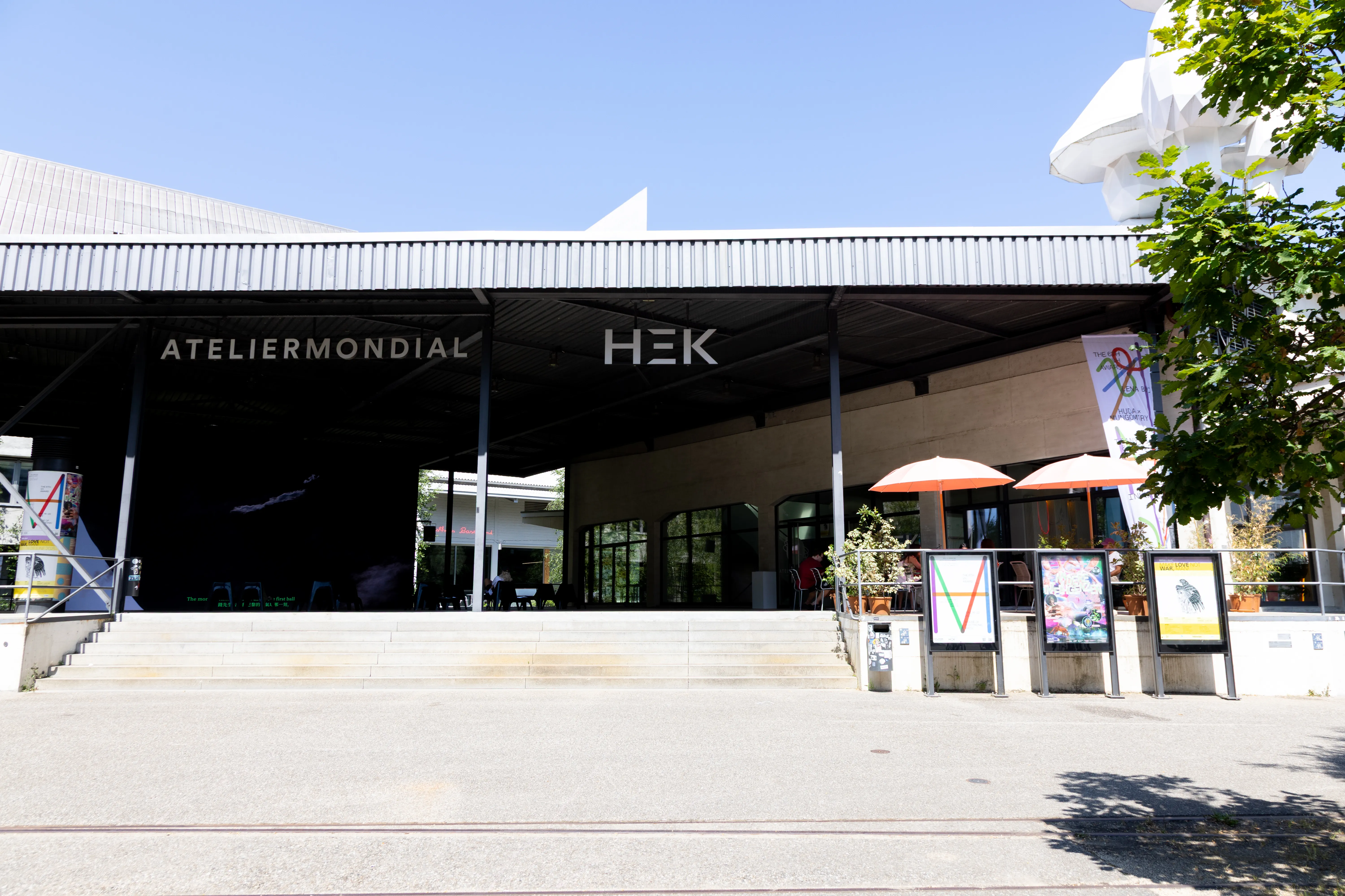 Widok z zewnątrz na fasadę budynku HEK – House of Electronic Arts z oznakowaniem ATELIERMONDIAL i HEK, szerokimi betonowymi schodami prowadzącymi do wejścia oraz miejscami do siedzenia na zewnątrz z parasolami.