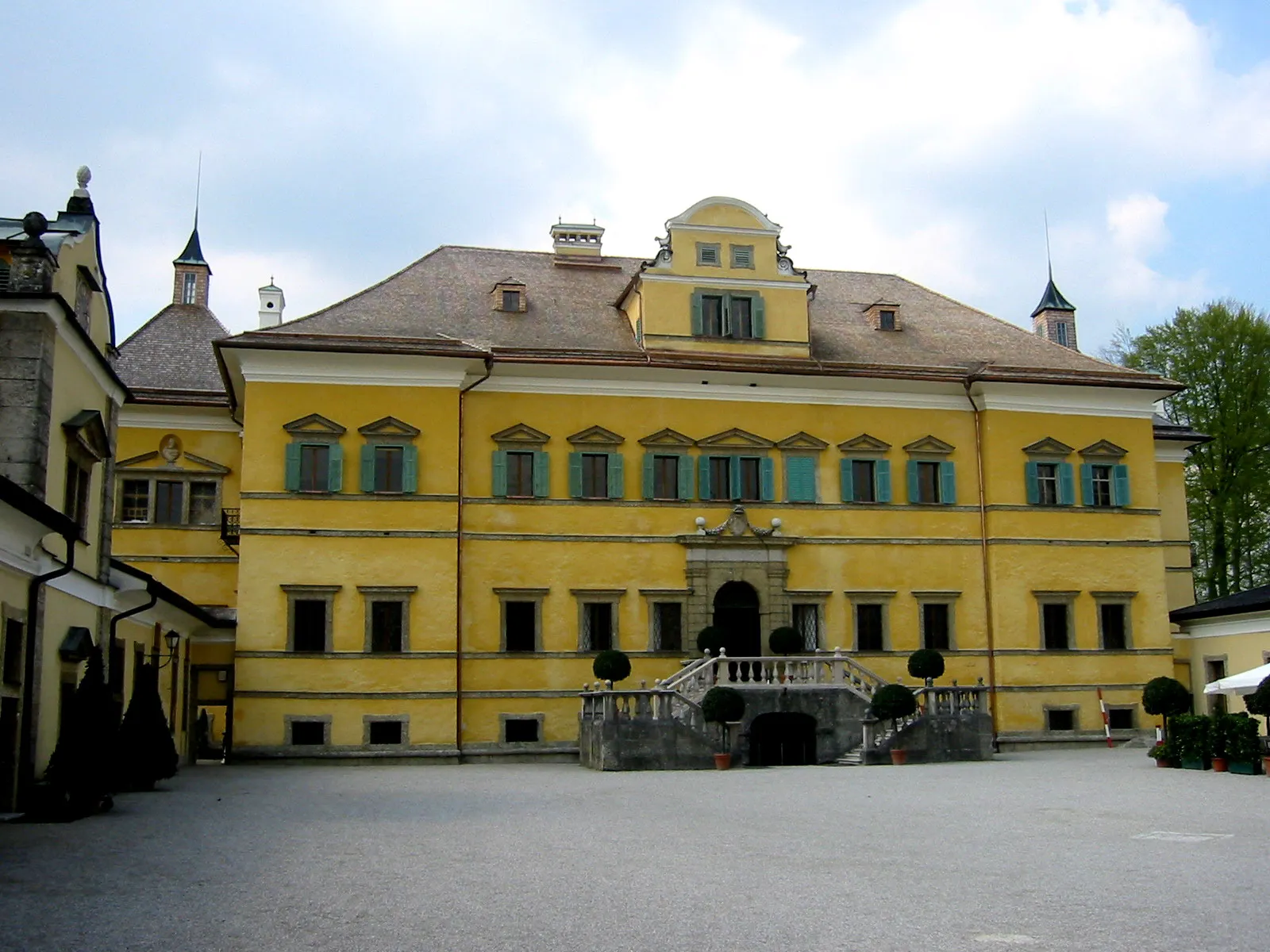 Fachada amarilla del Palacio de Hellbrunn con contraventanas verdes, escalera central y un patio pavimentado en el frente