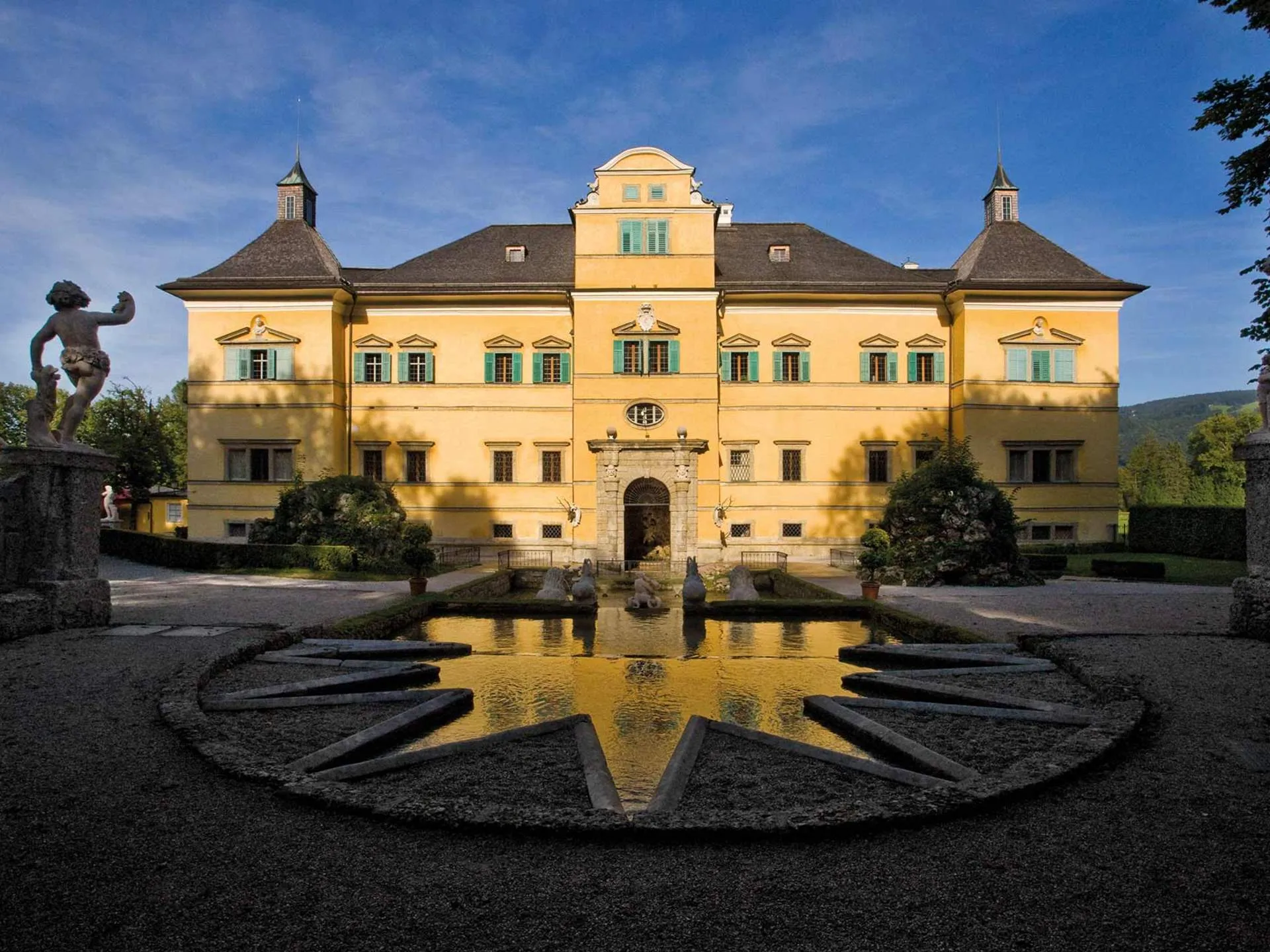 Fachada de un palacio barroco amarillo con contraventanas verdes reflejada en una fuente con forma de reloj de sol, con una estatua de bronce a la izquierda bajo un cielo azul.