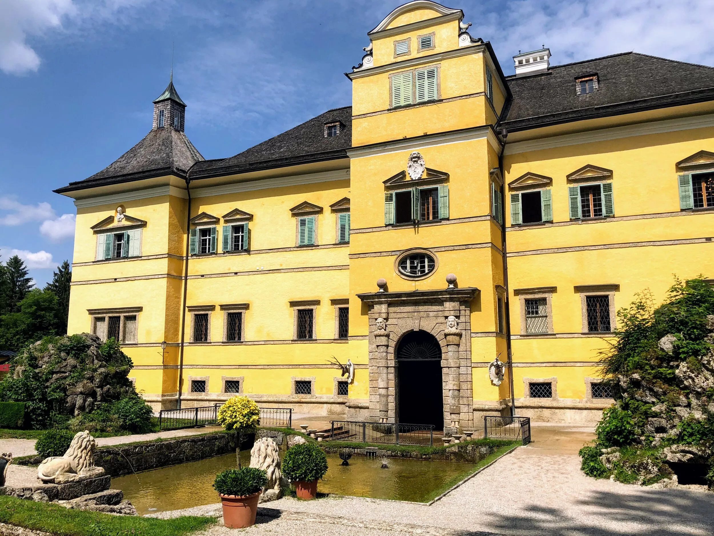 Fachada amarilla del Palacio de Hellbrunn con contraventanas verdes, entrada de arco de piedra y un pequeño estanque con plantas en macetas en primer plano