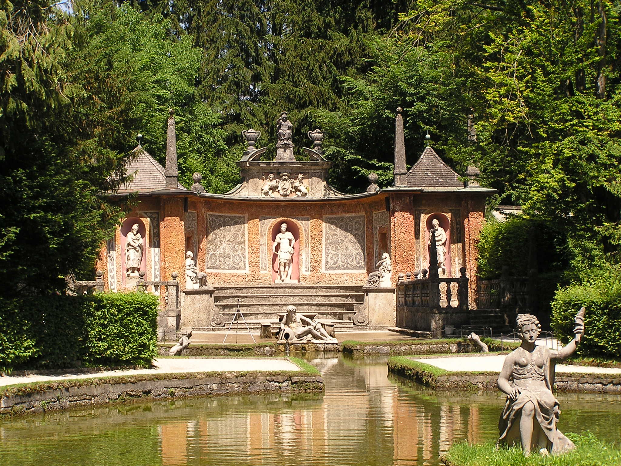Ornado complejo de fuentes barrocas con estatuas blancas en hornacinas, un estanque reflectante y una estatua sentada en primer plano, rodeado de frondosos árboles en el Palacio de Hellbrunn