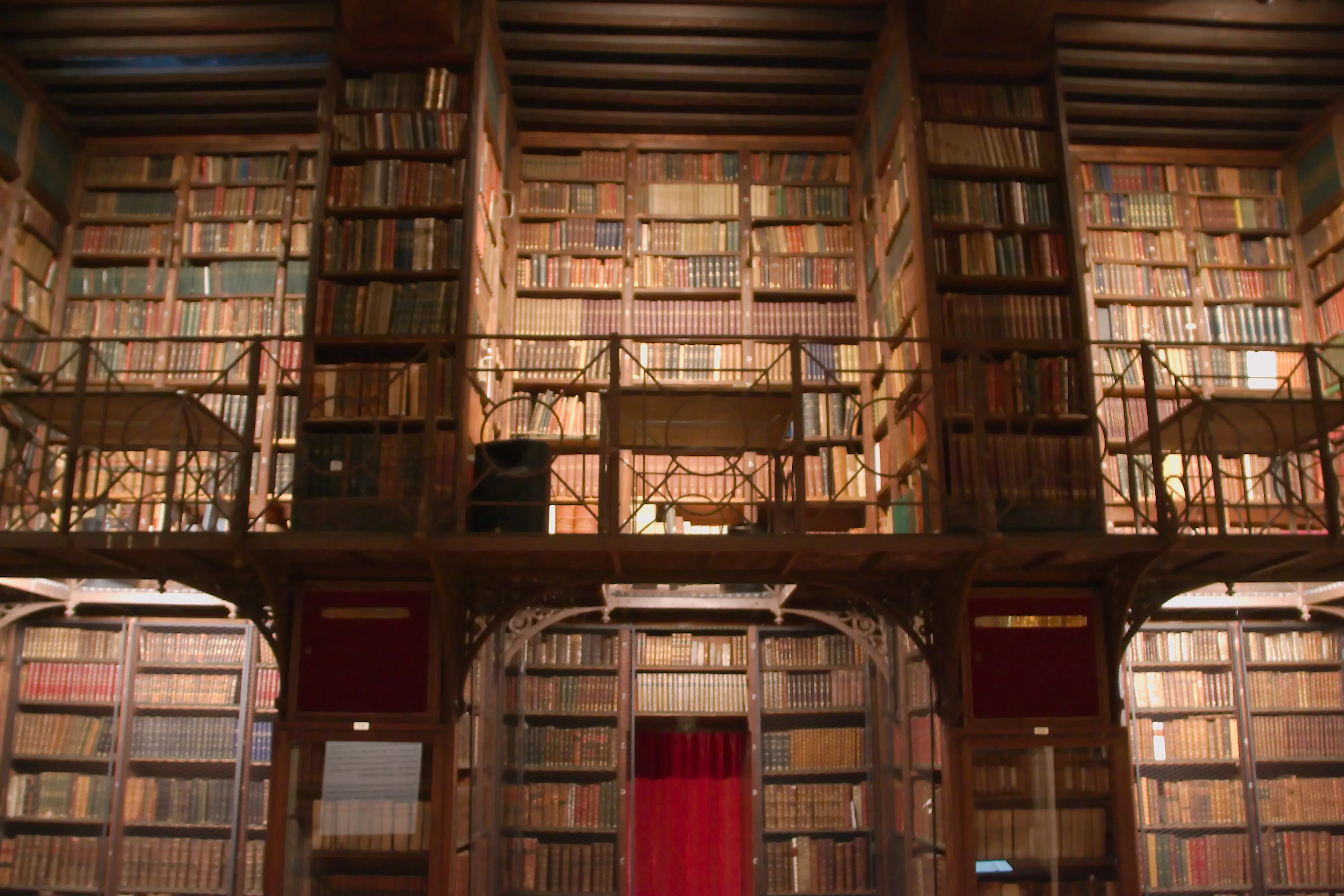 Interior de una biblioteca histórica con estanterías de madera de suelo a techo llenas de libros antiguos, una galería entresuelo con barandillas de hierro y una cortina roja en el centro.