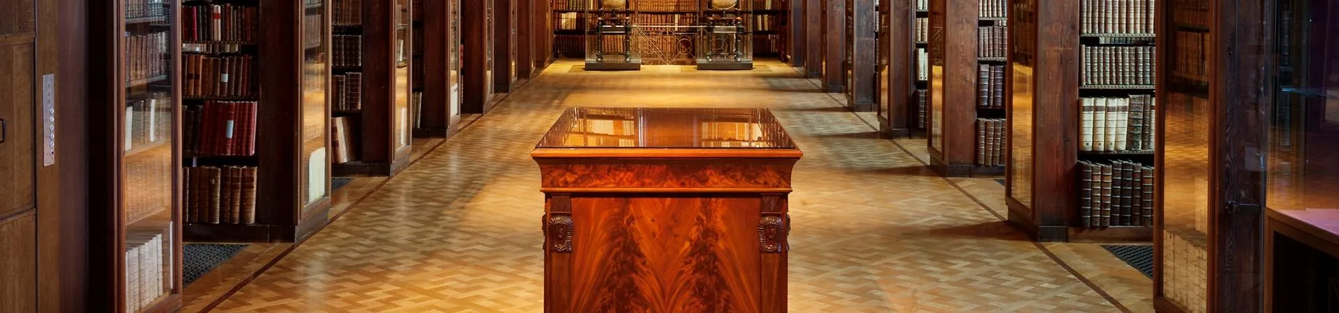 Larga sala de biblioteca con estanterías de madera altas a ambos lados, una vitrina central de madera y suelo de baldosas estampadas bajo una iluminación cálida