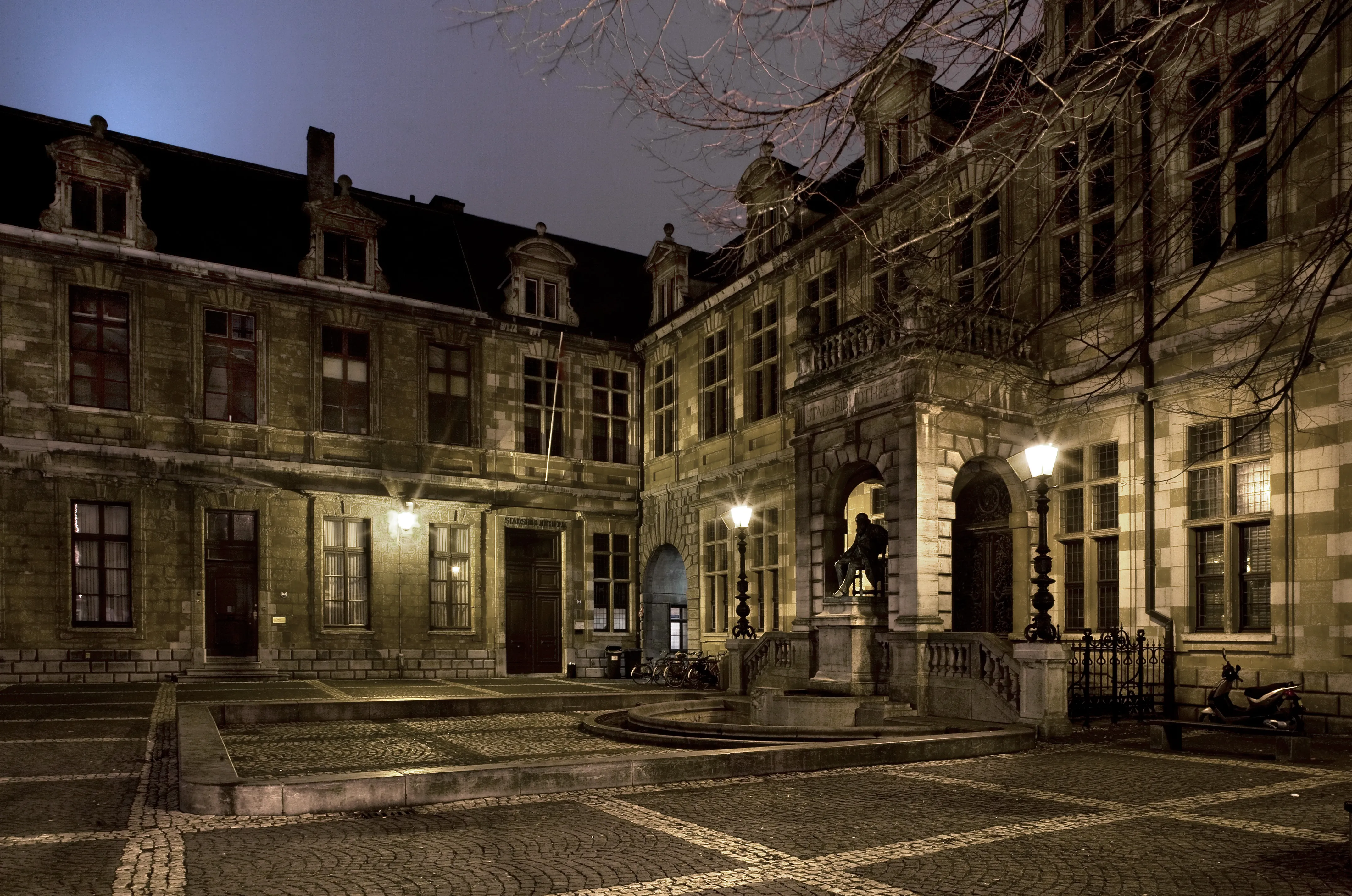 Vista nocturna del histórico patio de piedra de la Hendrik Conscience Heritage Library, con entradas arqueadas, una estatua central sobre un pedestal y pavimento de adoquines iluminado por farolas.
