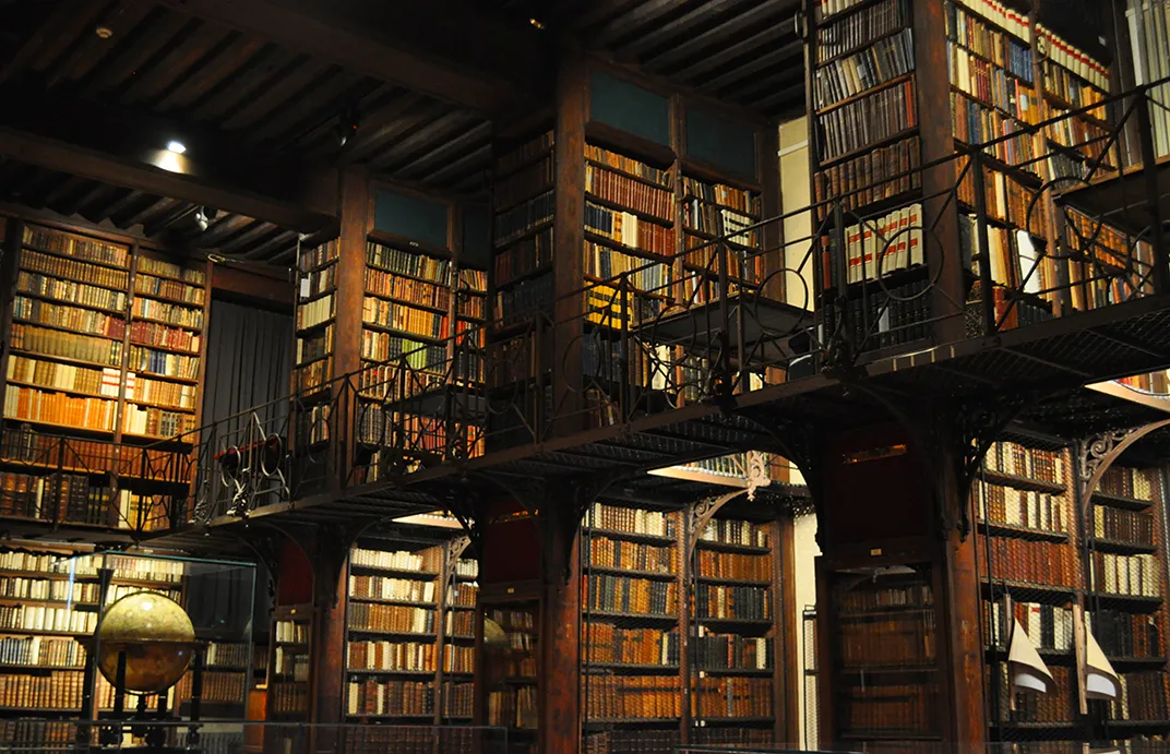 Wielopoziomowe wnętrze zabytkowej biblioteki z wysokimi drewnianymi regałami wypełnionymi starymi książkami, ciemnymi drewnianymi belkami, żelaznymi balustradami na balkonach oraz globusem na parterze