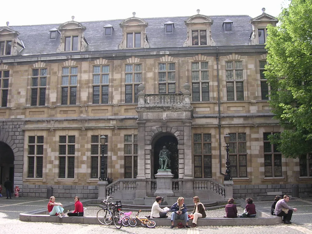 Fachada de piedra de la Hendrik Conscience Heritage Library con una estatua en una hornacina central arqueada, personas sentadas en un muro bajo en el patio y bicicletas aparcadas cerca