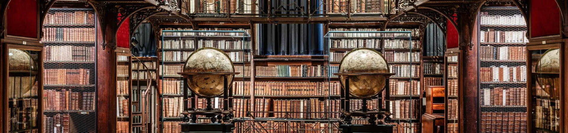 Vista panorámica de una sala de biblioteca histórica que cuenta con altas estanterías de madera llenas de libros y dos grandes globos terráqueos antiguos sobre soportes.
