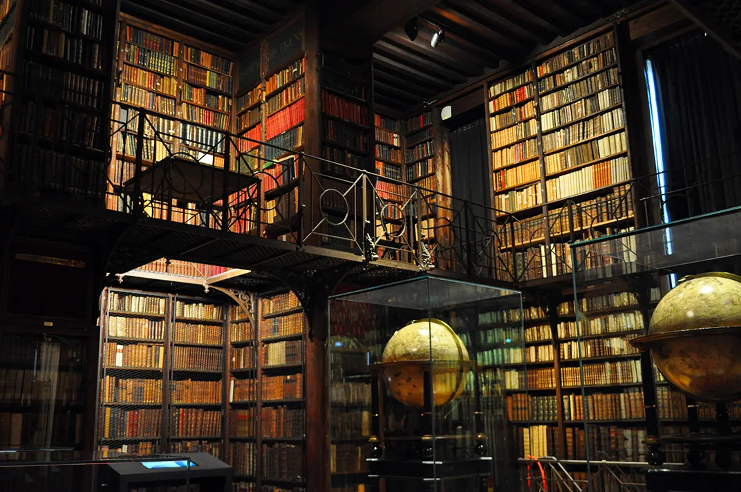 Interior de una biblioteca histórica con estanterías de madera altas llenas de libros antiguos, un balcón entresuelo con barandilla de hierro y dos globos terráqueos grandes en vitrinas de cristal.