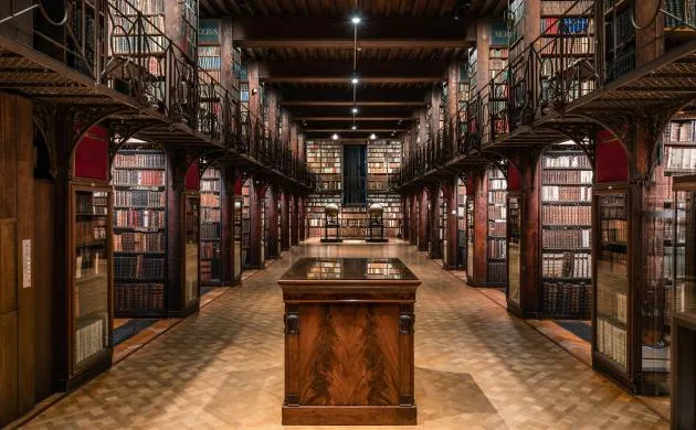 Vista interior alargada de la Sala Nottebohm con altas estanterías de madera a ambos lados, una mesa de exposición central de madera y suelo de baldosas bajo una iluminación cálida
