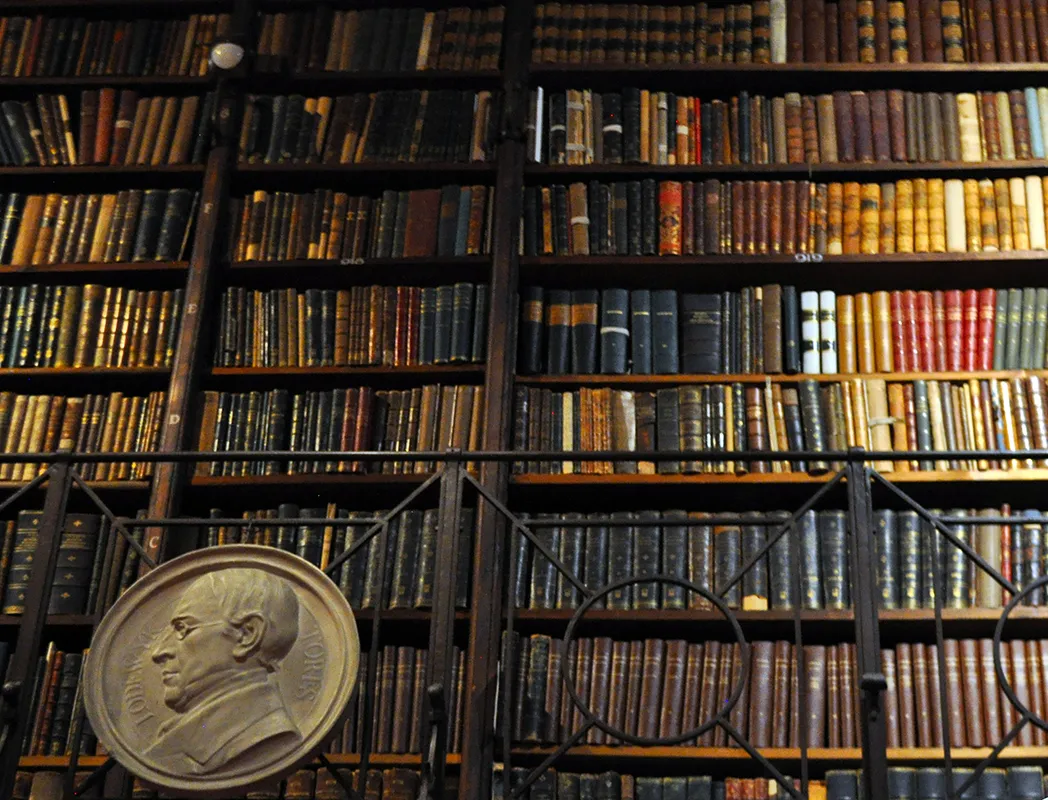 Altas estanterías de madera llenas de libros antiguos encuadernados en cuero en una biblioteca histórica, con un gran medallón de bronce en relieve sobre una balaustrada de hierro en primer plano