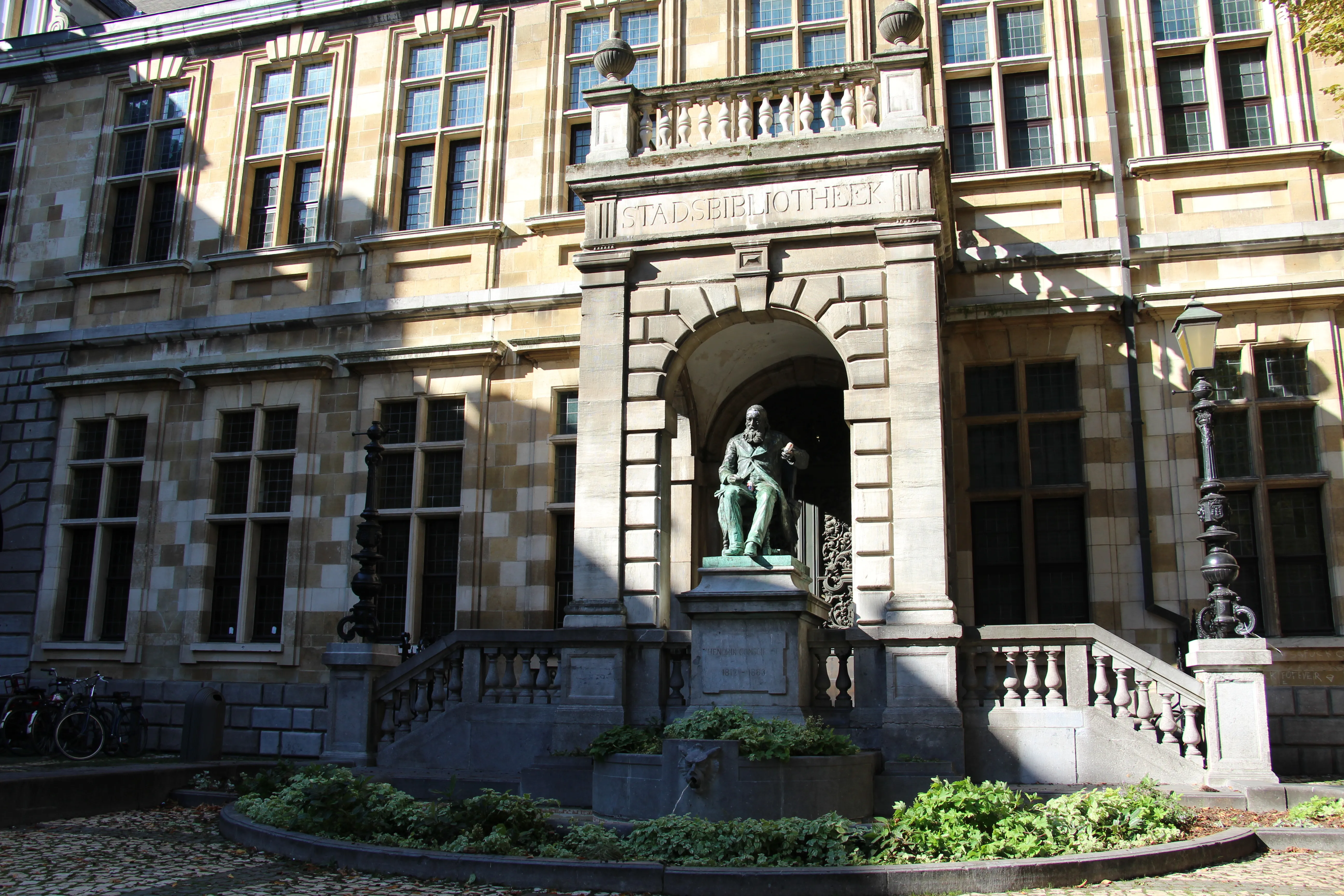 Kamienna fasada Biblioteki Dziedzictwa Hendrika Conscience'a z centralnym łukowym wejściem z posągiem, dużymi oknami wielodzielnymi, kamiennymi balustradami oraz brukowanym dziedzińcem z klombem