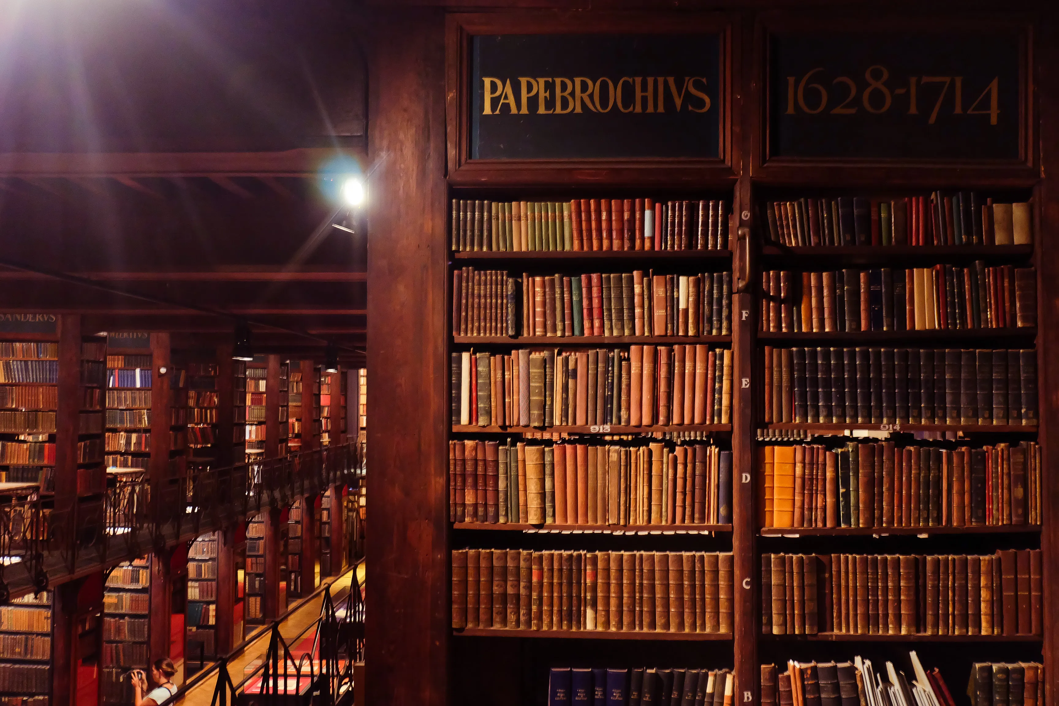 Wysokie, ciemne drewniane regały wypełnione zabytkowymi książkami w zabytkowej bibliotece, z napisem "PAPEBROCHIUS 1628–1714" nad półkami i ciepłym oświetleniem punktowym