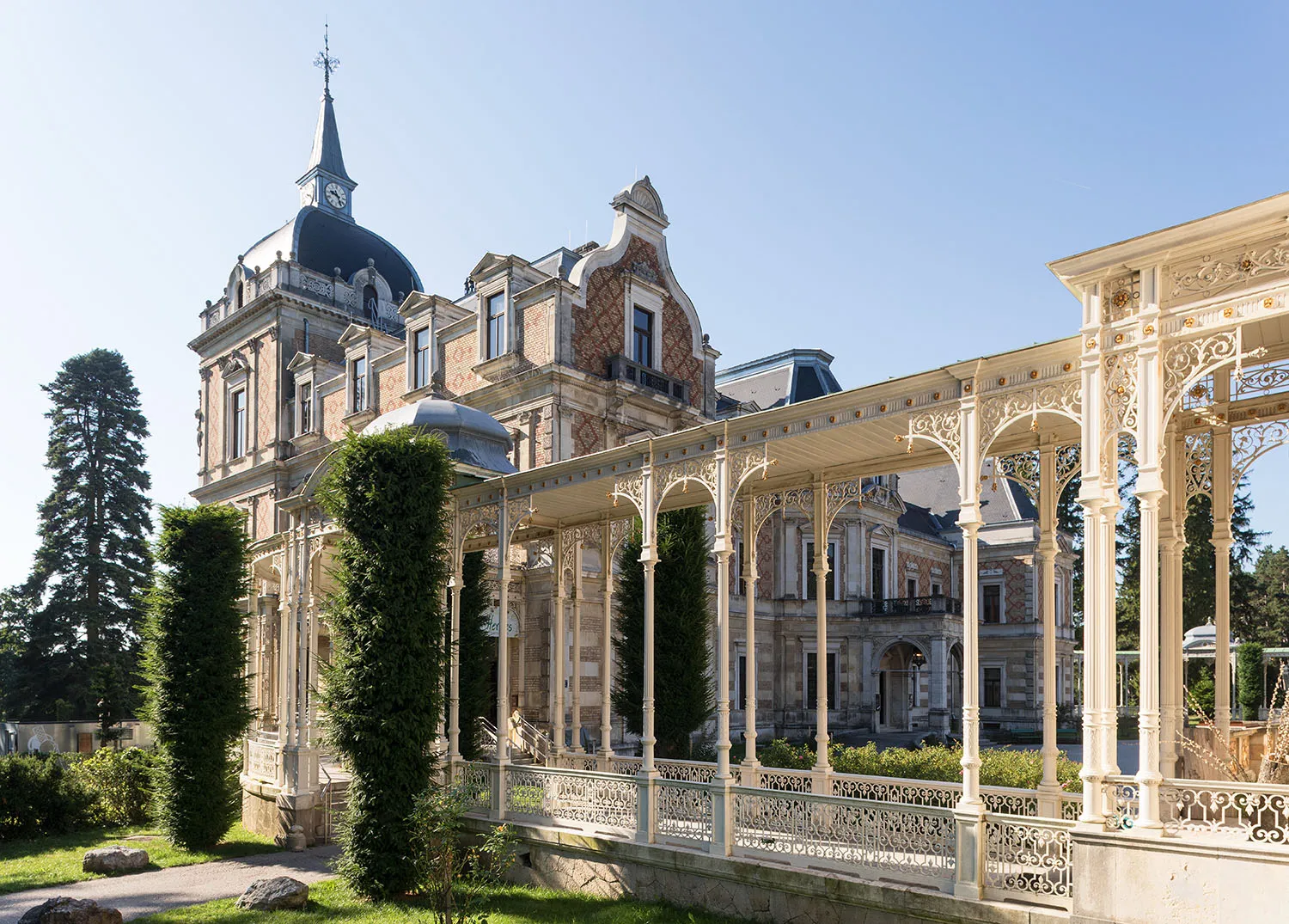 A grand historic villa with a blue-domed spire and ornate white iron colonnade under a clear blue sky
