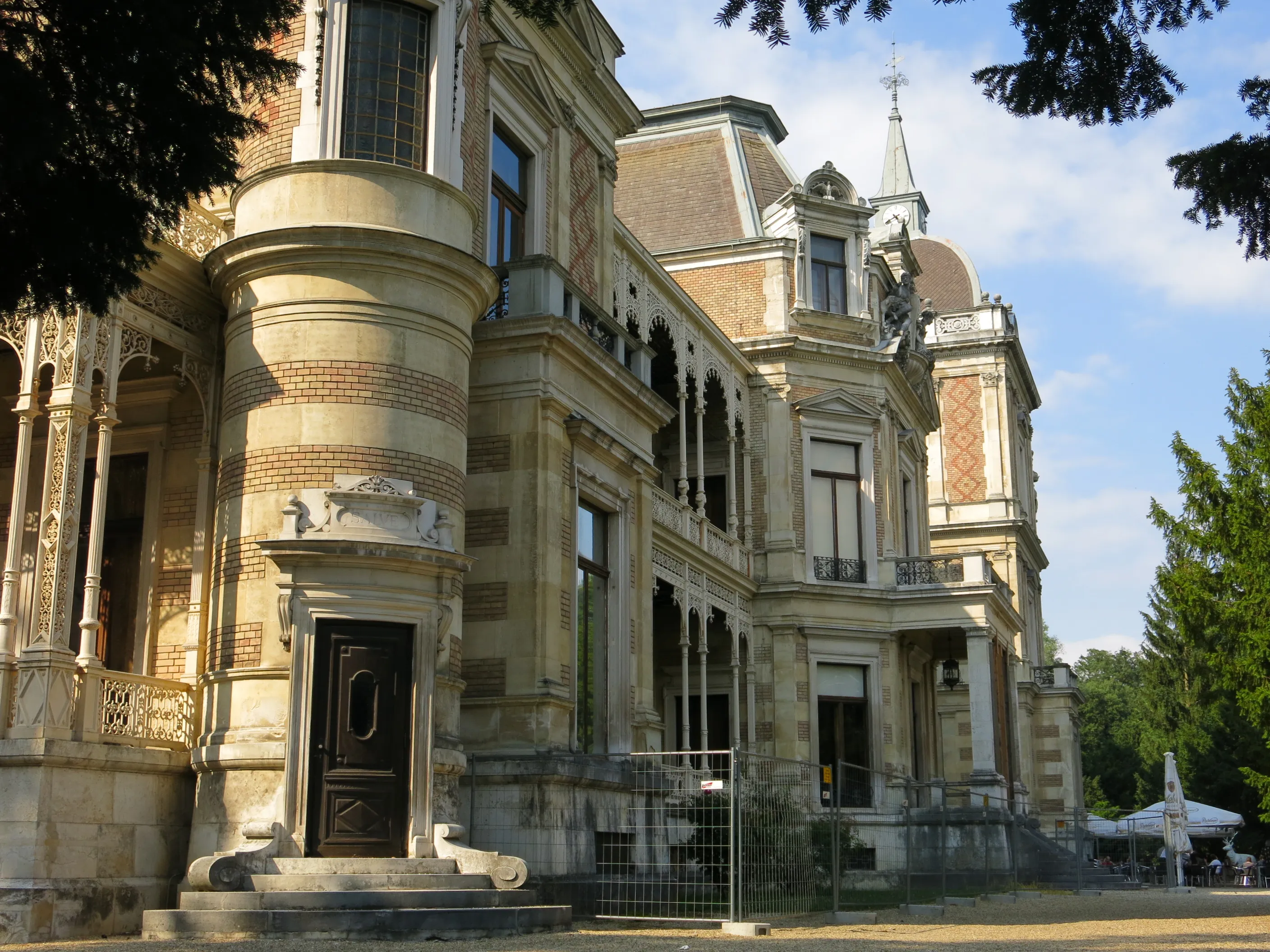 Zewnętrzna część Hermesvilla, okazałej zabytkowej willi z ozdobną kamienną architekturą, łukowymi oknami i dekoracyjnym wejściem, otoczonej drzewami i zielenią.