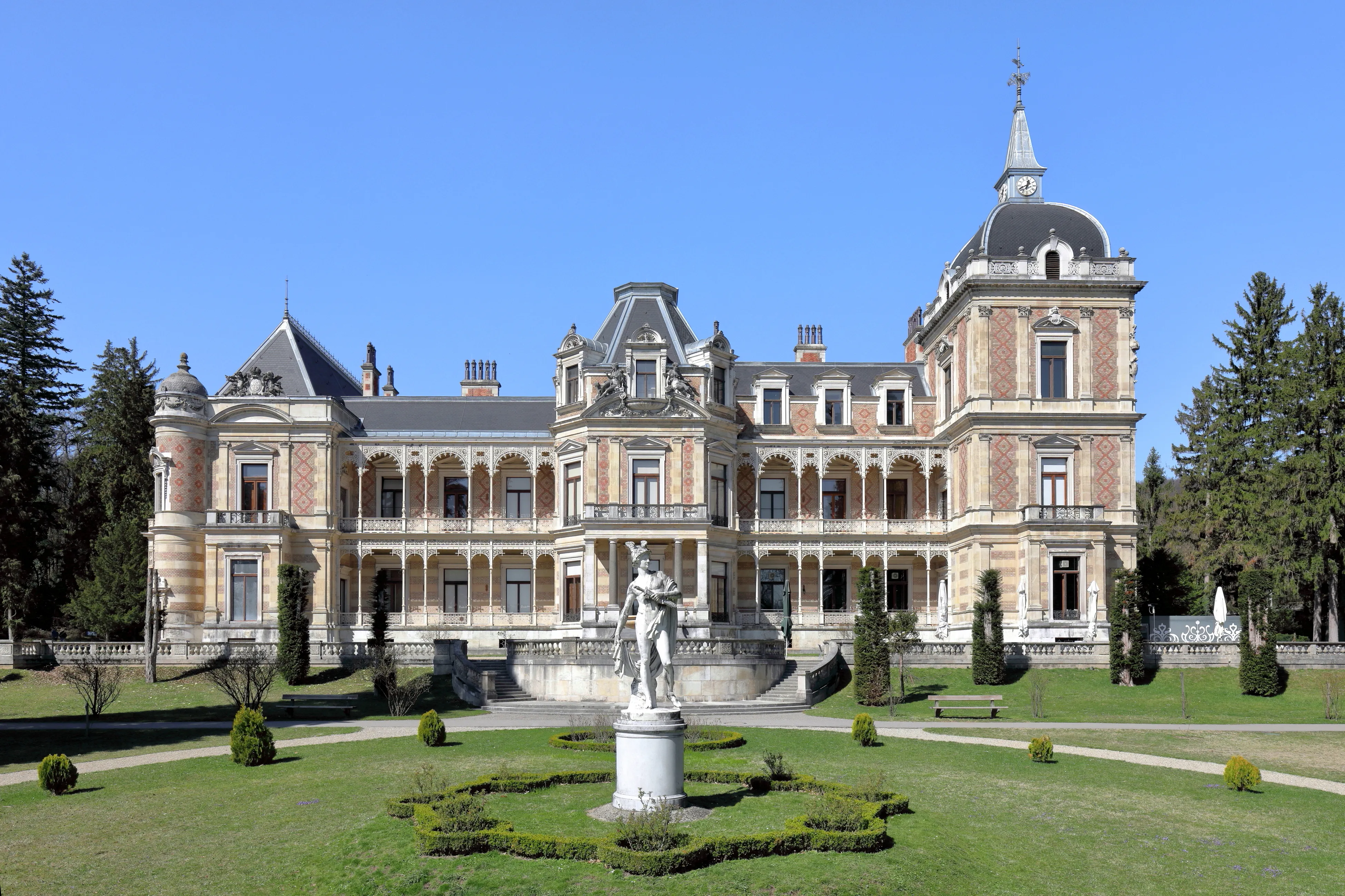 A large historic villa with a central dome and ornate architecture, surrounded by green lawns and trees under a blue sky.