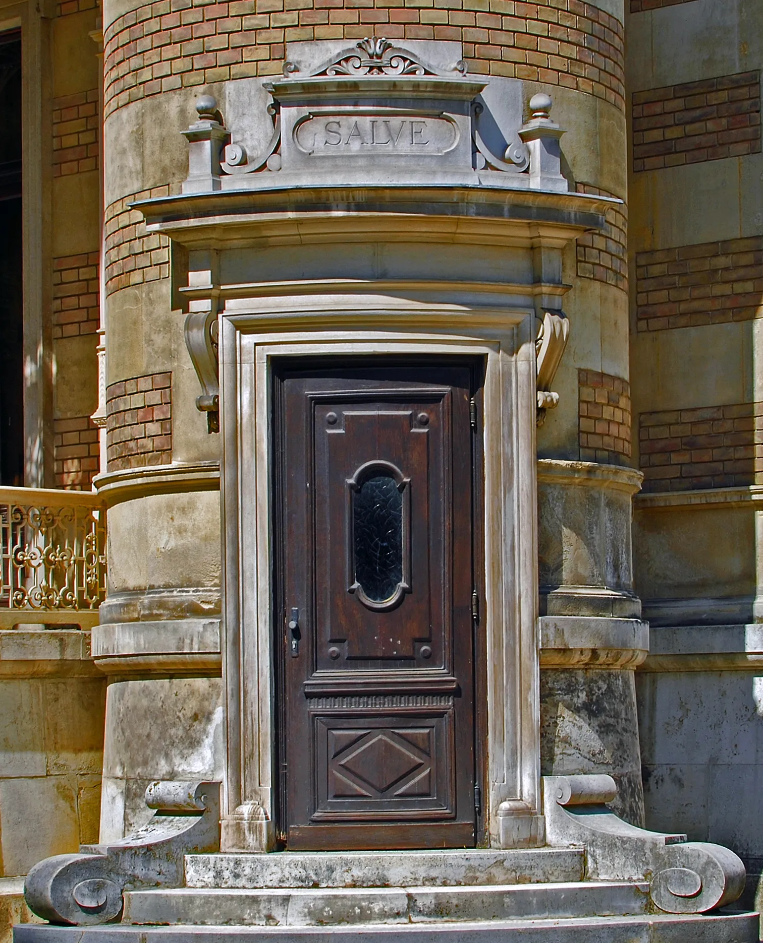 Close-up of a dark wooden door with an oval window, set within a stone archway with the inscription 'SALVE' above, on a brick and stone building facade.