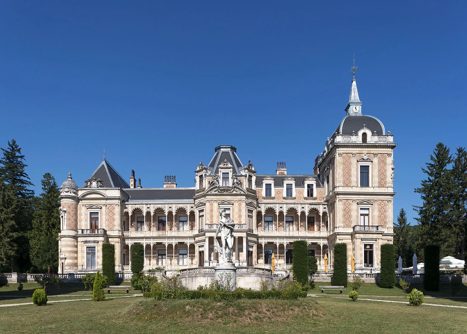 Large historic villa with ornate architecture, a central fountain, and manicured gardens under a clear blue sky