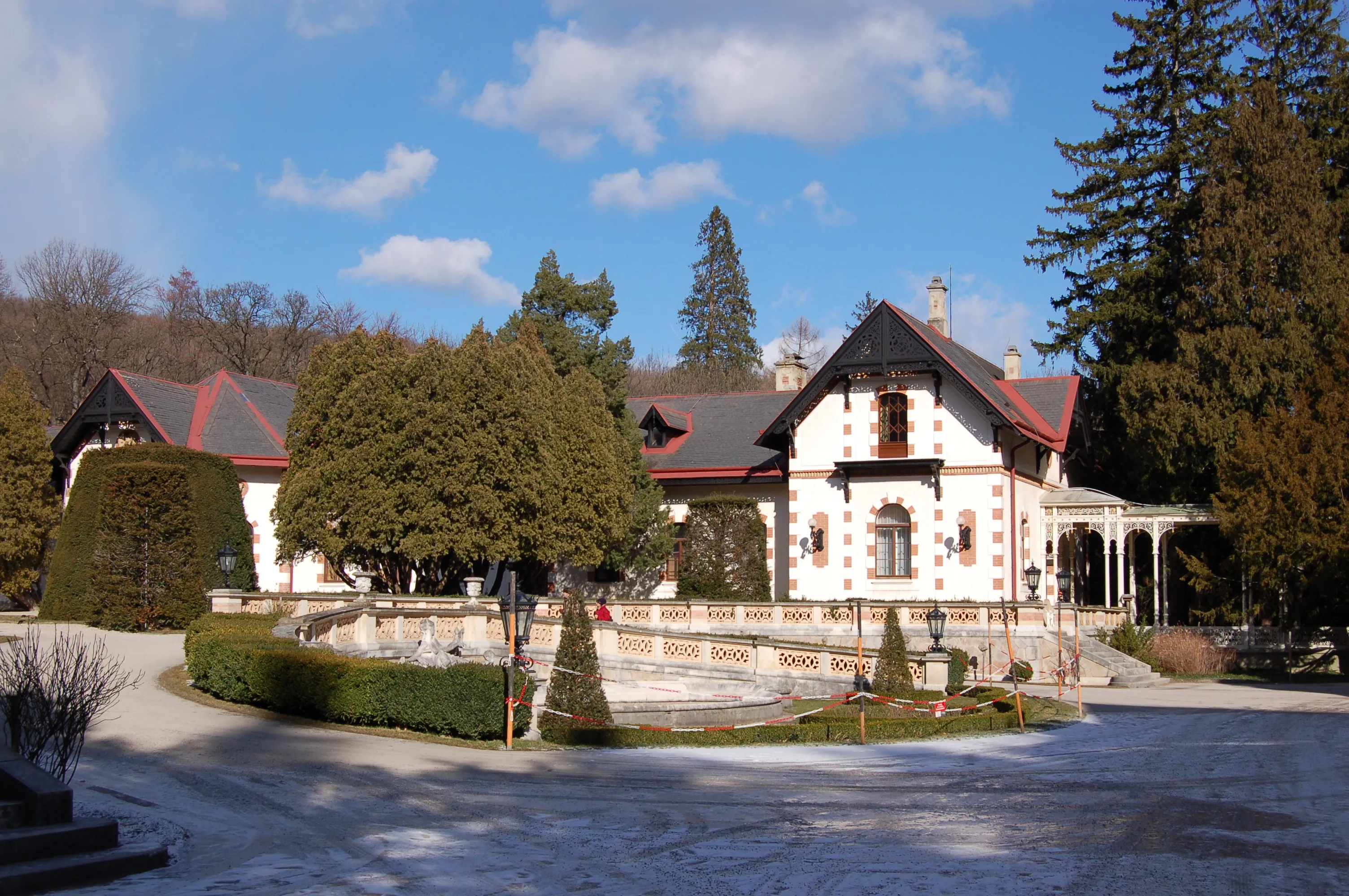 The Hermesvilla, a white building with red trim and a dark roof, surrounded by trees and manicured hedges under a blue sky.