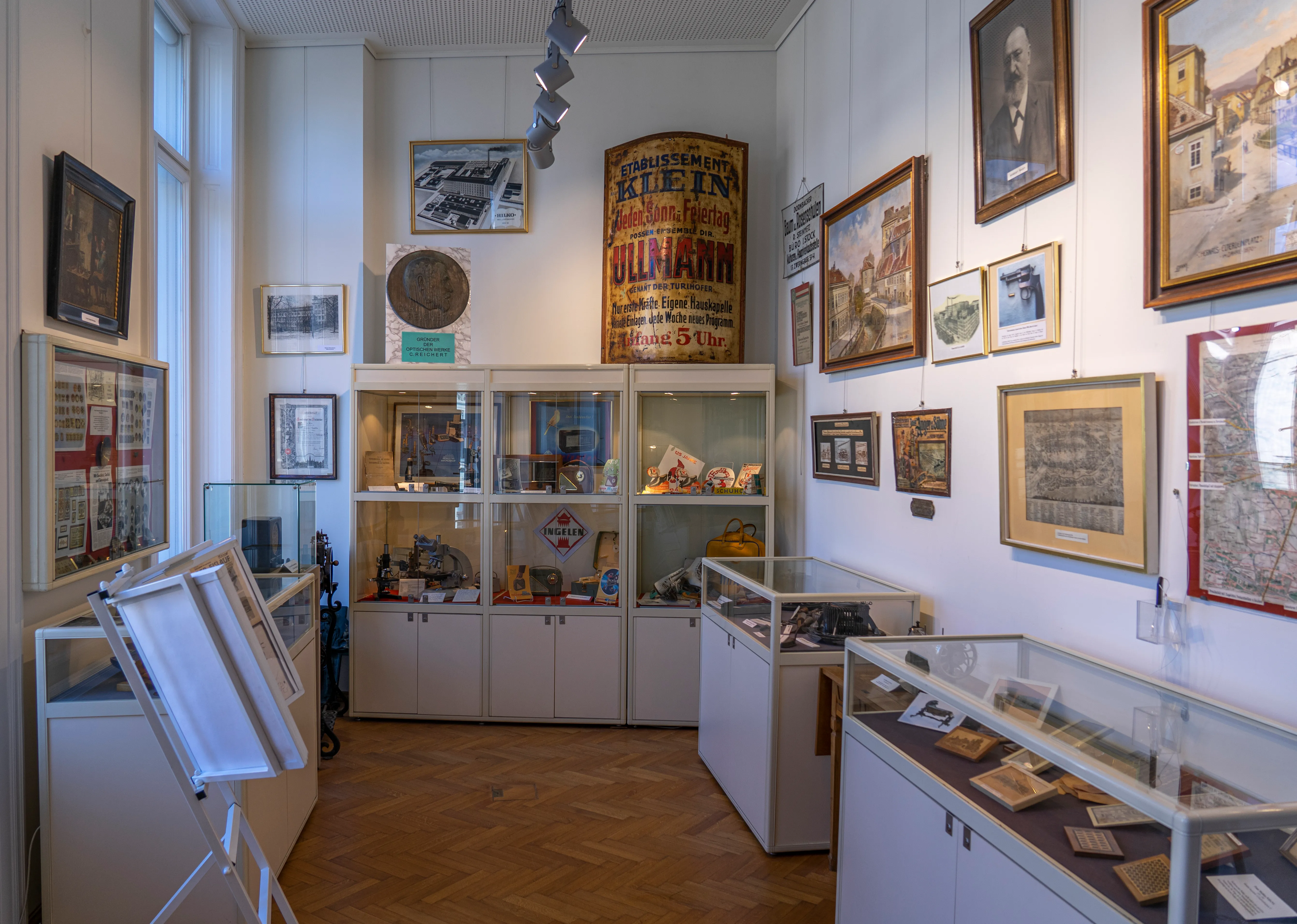 Interieur van een museum met witte vitrines, ingelijste foto's aan de muren en een grote poster in het midden