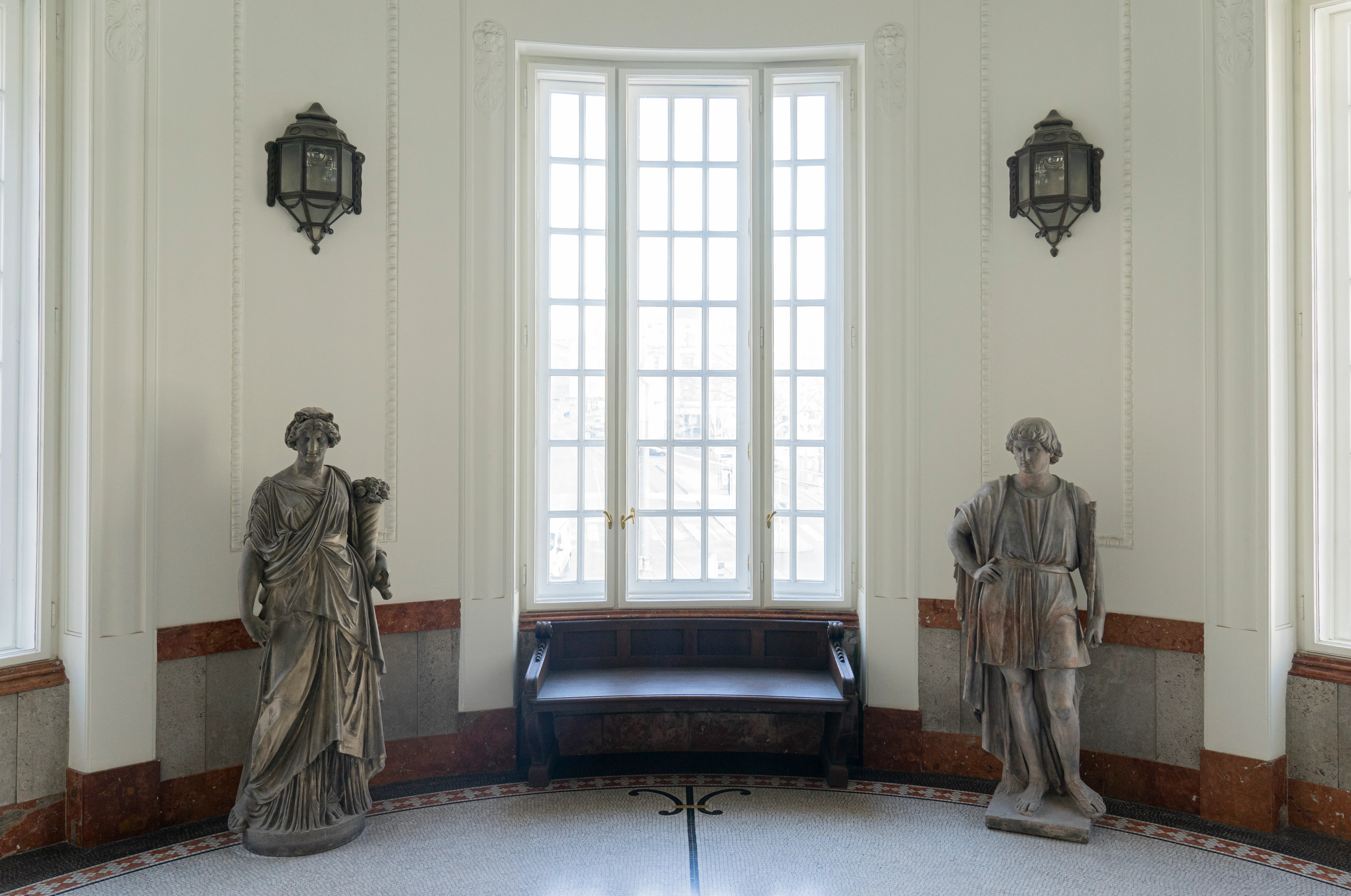 Twee klassieke beelden staan aan weerszijden van een groot raam in een museumgang met witte muren en een betegelde vloer.