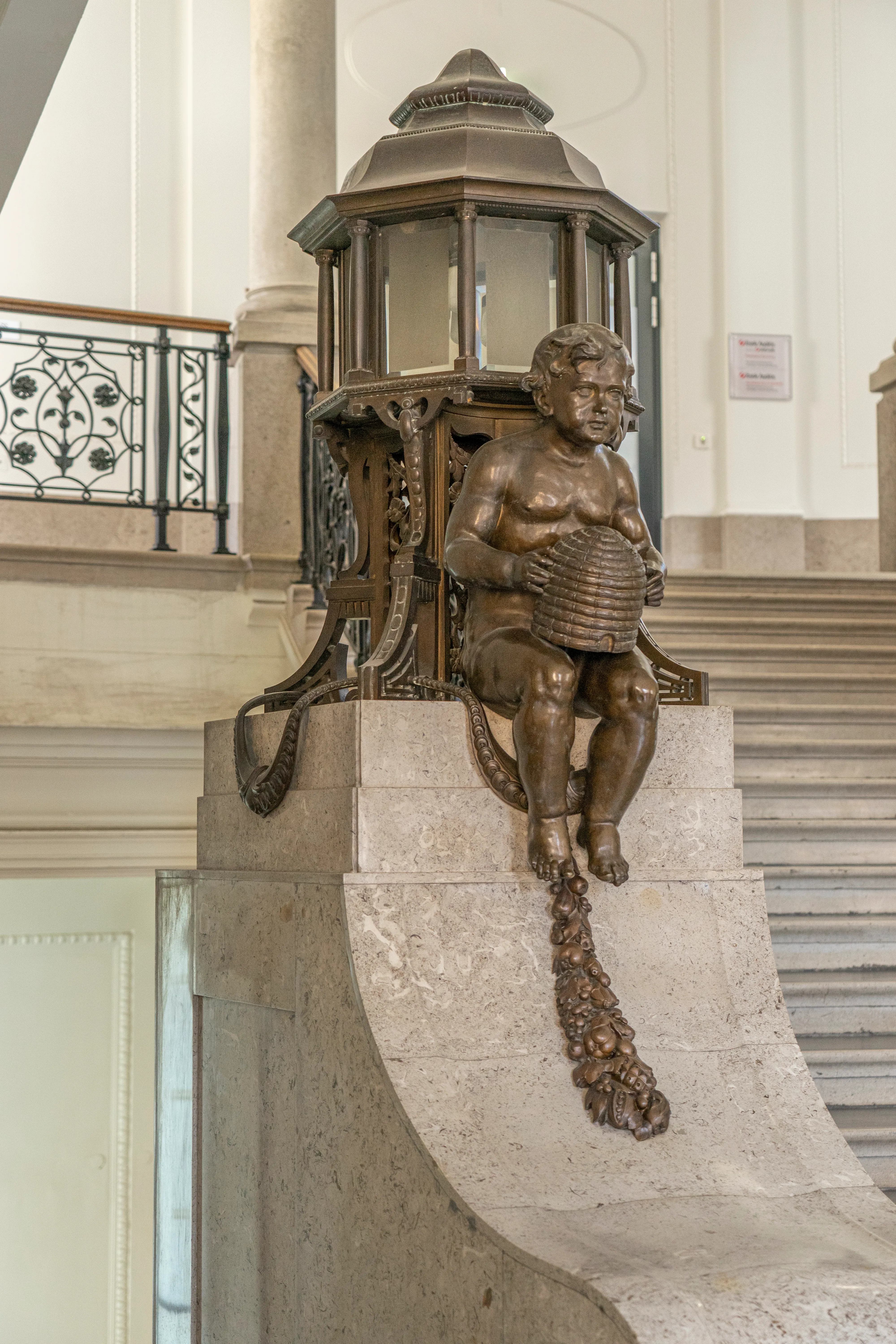 Bronzen beeld van een cherubijn met een mand op een stenen sokkel in een museuminterieur