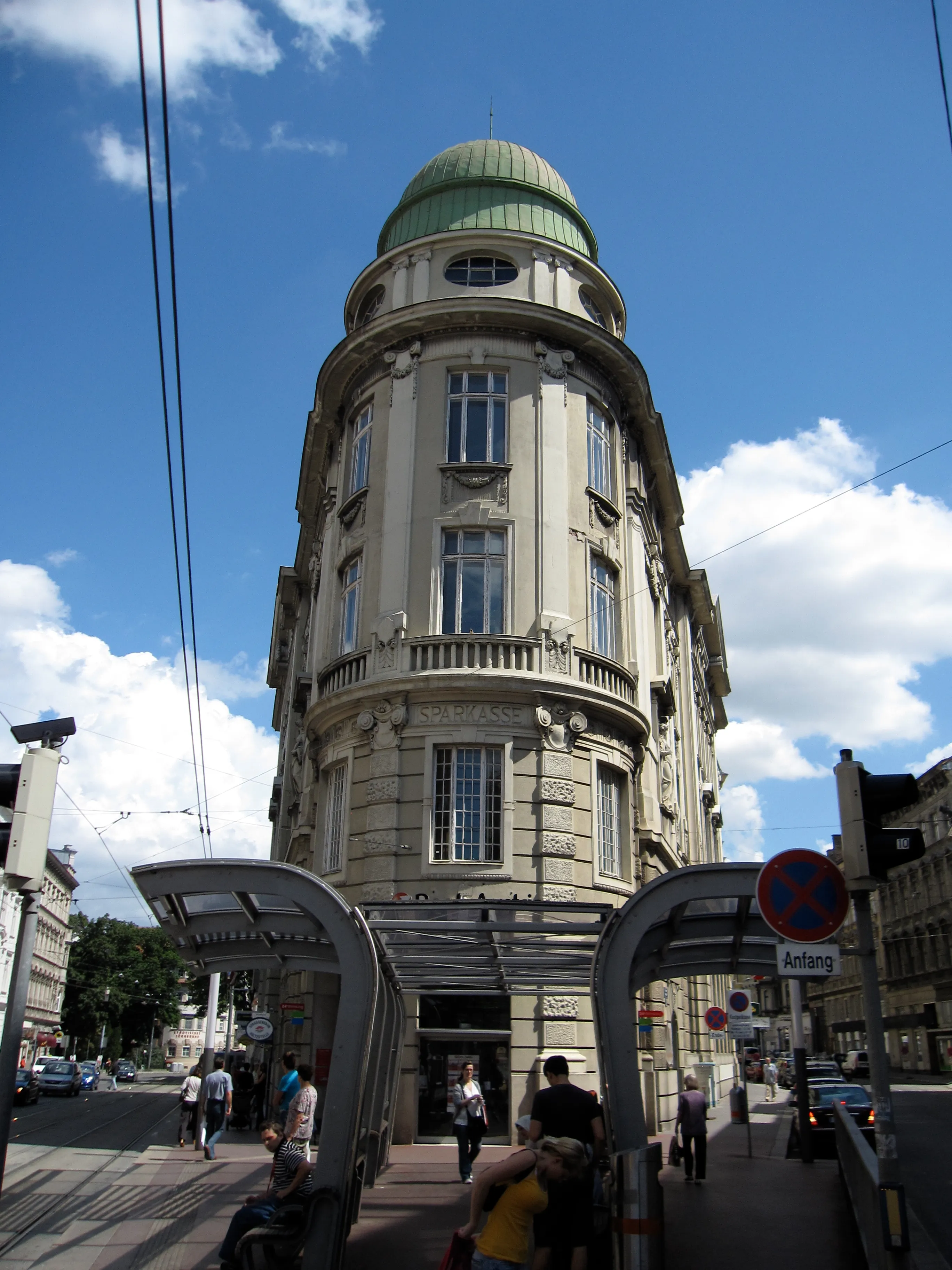 Un edificio alto con una cúpula verde y arquitectura clásica, ubicado en una esquina de la calle con marquesinas de tranvía y gente caminando cerca.