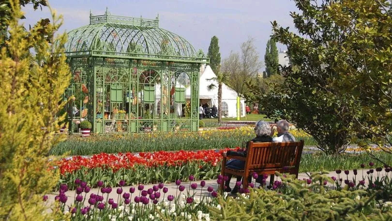 Green glasshouse surrounded by red and purple tulips with people sitting on a bench in the foreground
