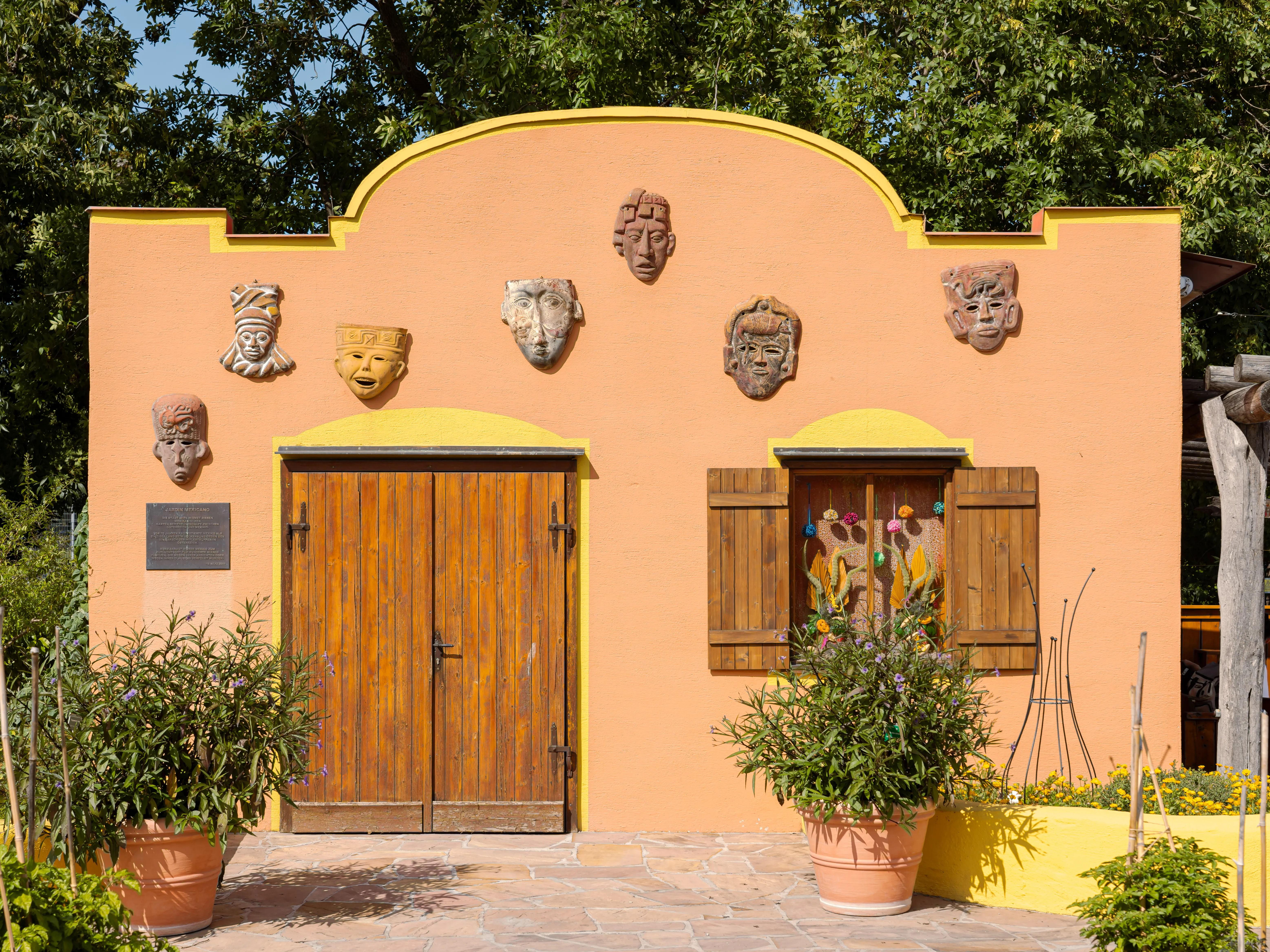 Orange building facade with yellow trim, wooden doors, and decorative masks, surrounded by potted plants and trees