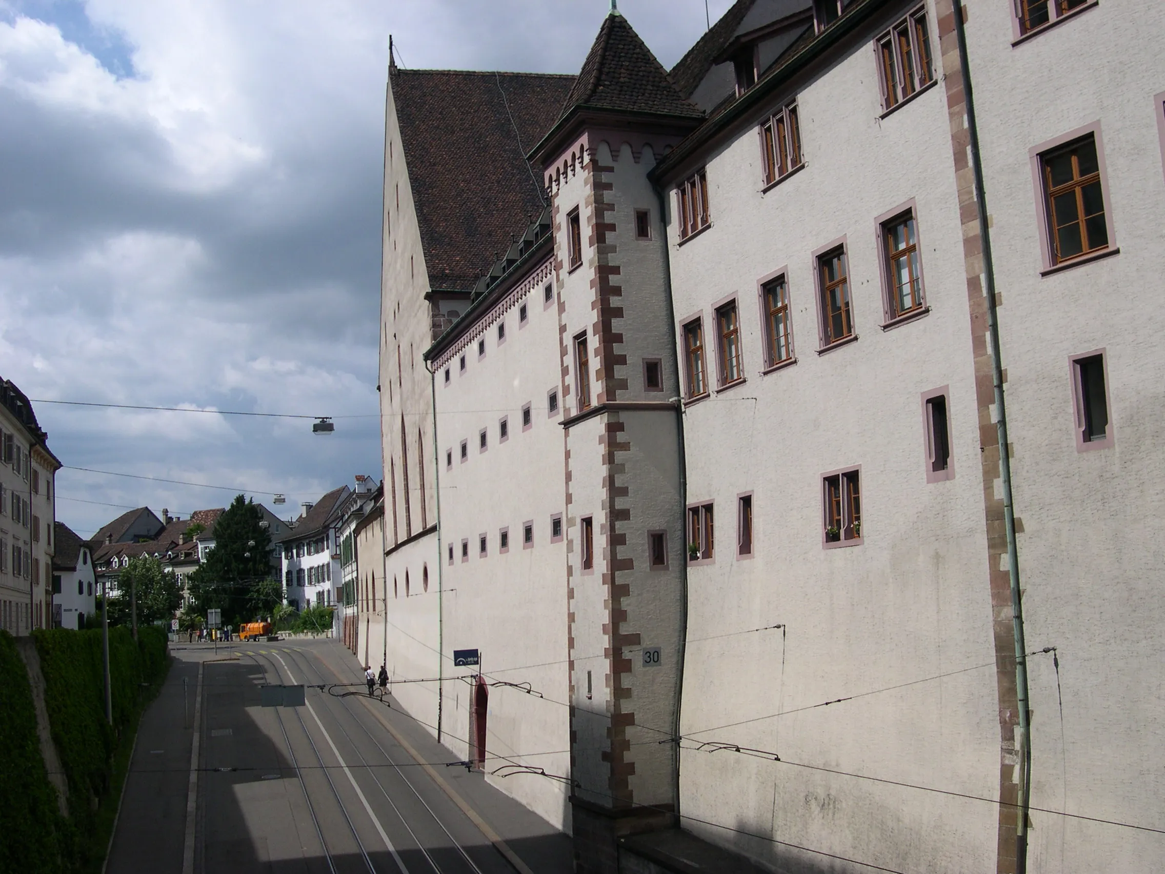 Wit gepleisterd historisch gebouw met een hoektoren en afwerking van rode zandsteen langs een straat met tramrails in Basel