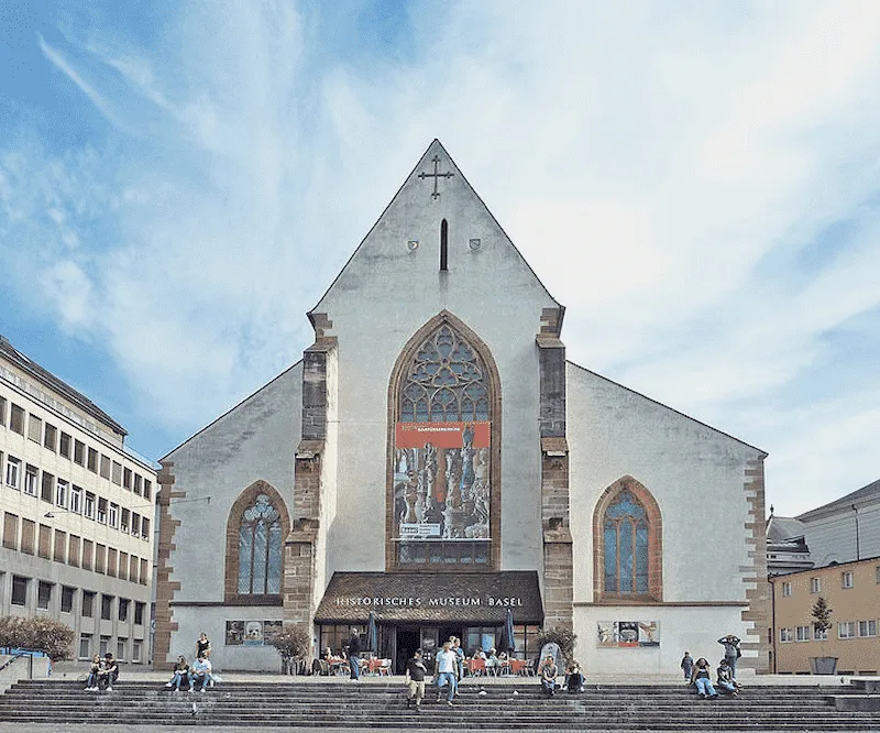 Buitenaanzicht van het Historisch Museum Bazel bij de Barfüsserkirche, met de witte kerkgevel met een grote centrale muurschildering, boogramen en mensen die op de voortrap zitten.