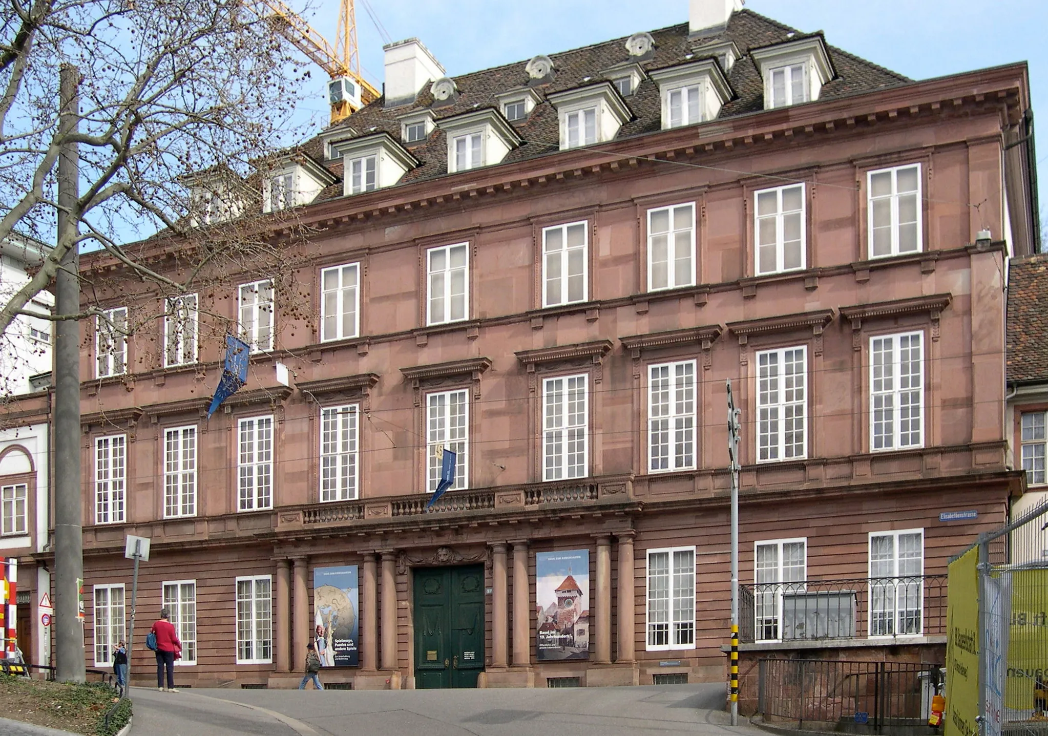 Groot roodbruin klassiek gevelgebouw met rijen wit omlijste ramen, een groene centrale toegangsdeur en blauwe vlaggen, met een paar voetgangers op het trottoir ervoor.