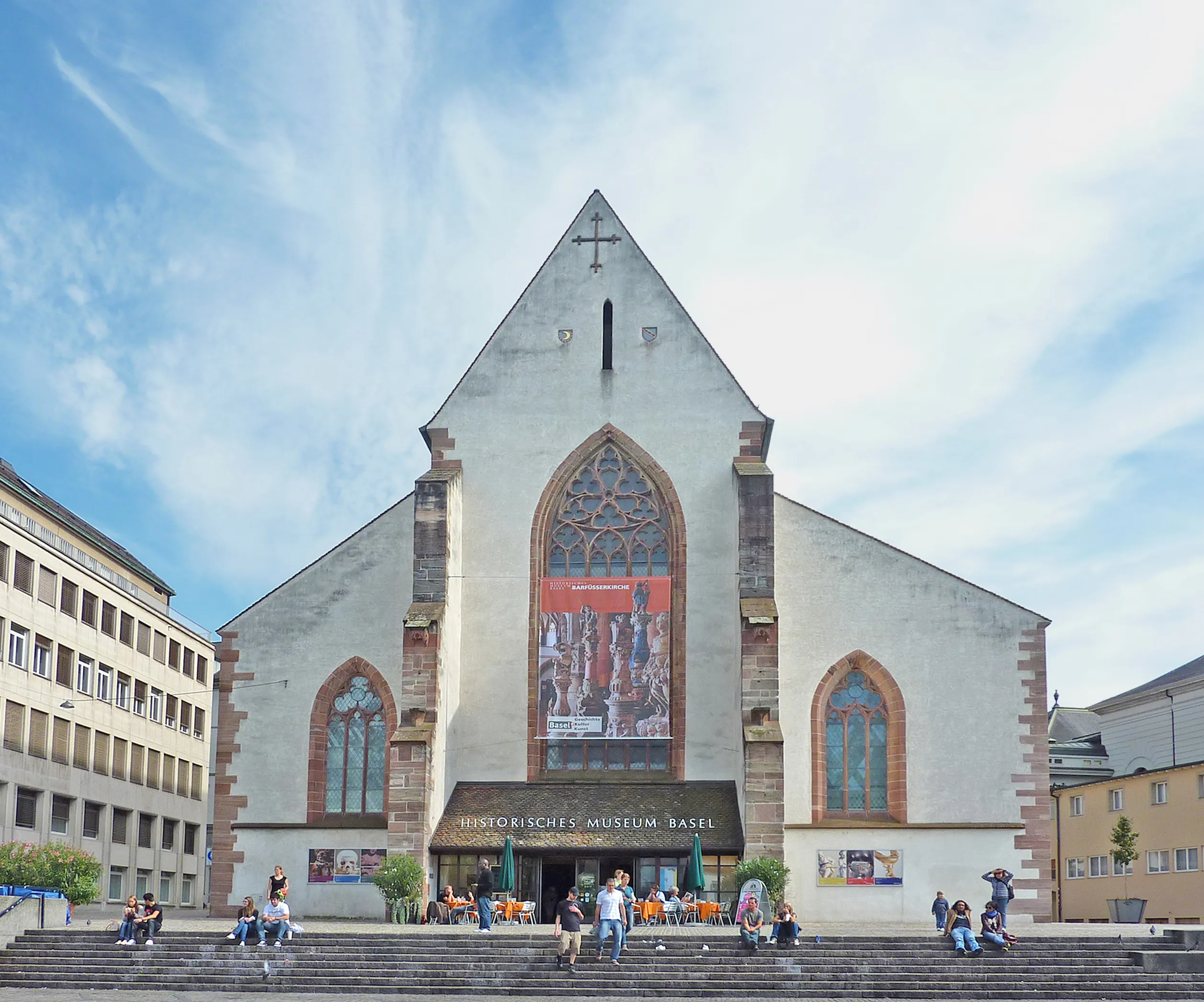 Buitenaanzicht van het Historisch Museum Bazel met de witte kerkgevel met gotische spitsboogvensters, een grote muurschildering als banner en mensen die op de trappen aan de voorzijde zitten.