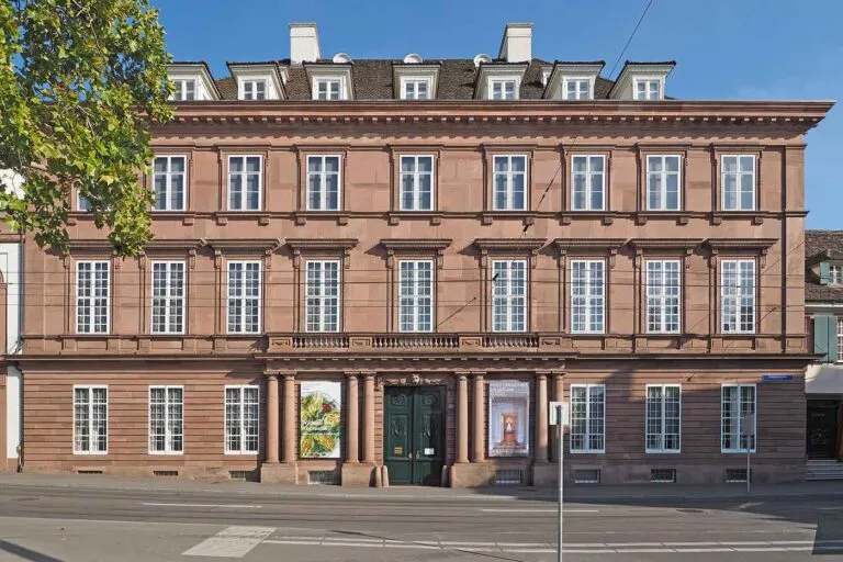 Rozeachtig rode historische gevel van een grachtenpand met meerdere rijen wit omrande ramen, een centrale groene voordeur en posters aan weerszijden van de ingang
