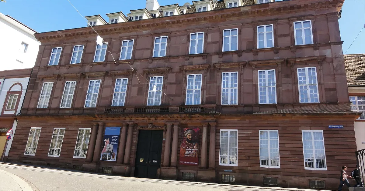 Rodbruine historische gevel van een herenhuis met meerdere verdiepingen, rijen wit omlijste ramen en een donkere voordeur geflankeerd door tentoonstellingsbanners