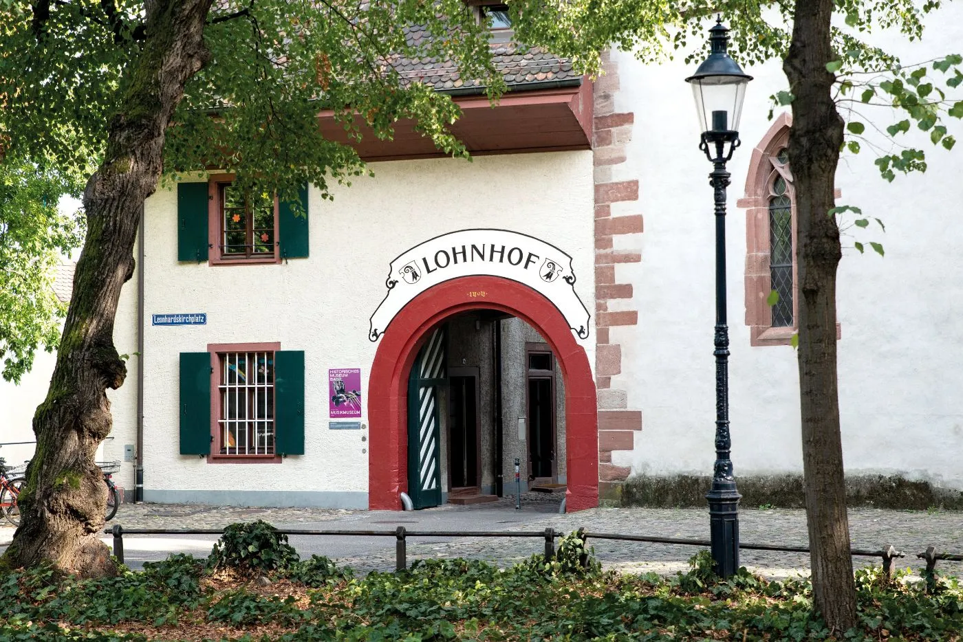 Fachada blanca con una entrada de arco rojo etiquetada como 'Lohnhof', contraventanas verdes y una farola negra enmarcada por árboles.