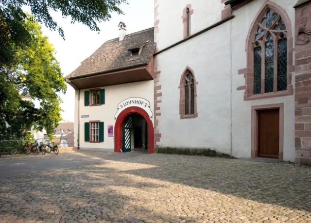 Edificio blanco con una entrada de arco rojo y ventanas góticas situado en un patio empedrado, con bicicletas aparcadas a un lado y árboles al fondo
