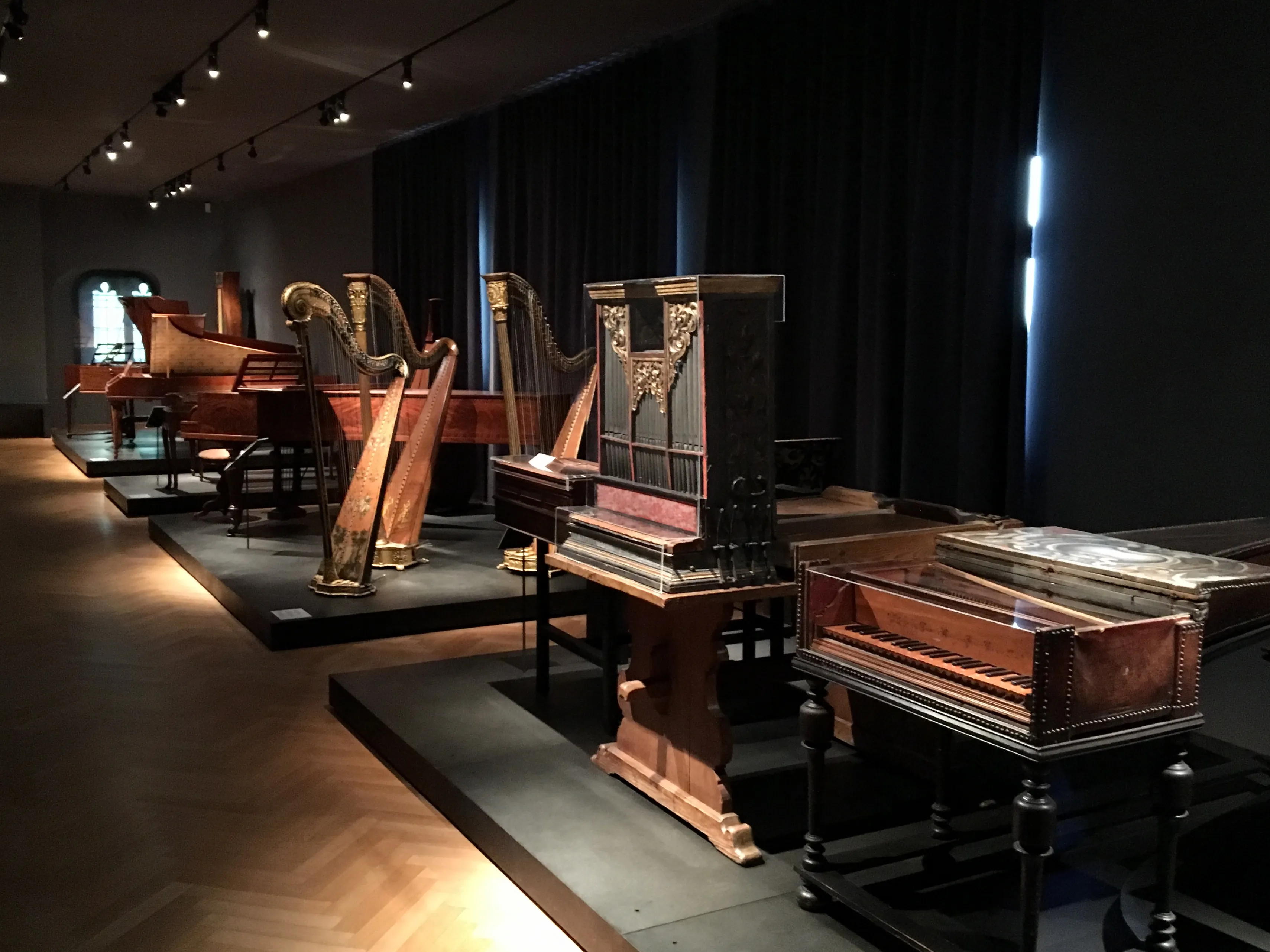 Instrumentos musicales históricos, incluyendo arpas de madera e instrumentos de teclado antiguos, expuestos sobre plataformas elevadas en una sala de museo con iluminación tenue y focos dirigidos.