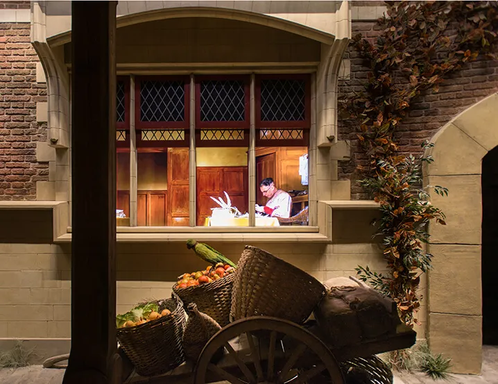 Escenario de exposición temática que muestra una escena de Brujas del siglo XV, con una figura trabajando en un escritorio visible a través de una ventana de vidrio emplomado, cestas de verduras y un carro de madera en primer plano, y hiedra trepando por una pared de ladrillo junto a un arco de piedra.