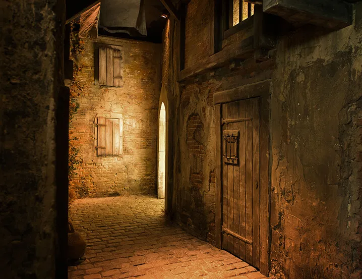 Callejón de adoquines con iluminación tenue, paredes de ladrillo envejecido, una puerta de madera con herrajes de hierro y ventanas con contraventanas en un entorno interior de temática medieval.