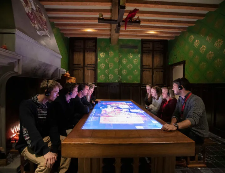 Grupo de personas con auriculares reunidas alrededor de una gran mesa táctil interactiva en una sala temática con papel pintado verde estampado, techo con vigas de madera y una chimenea.