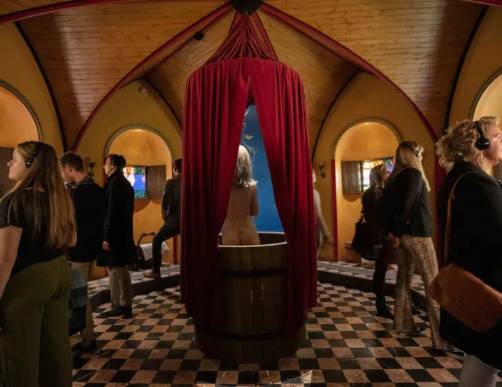 Interior del Historium de Brujas donde se muestran visitantes con auriculares de audio reunidos alrededor de un barril de madera central cubierto con cortinas rojas, situado en una sala abovedada con arcos y un suelo de damero blanco y negro.