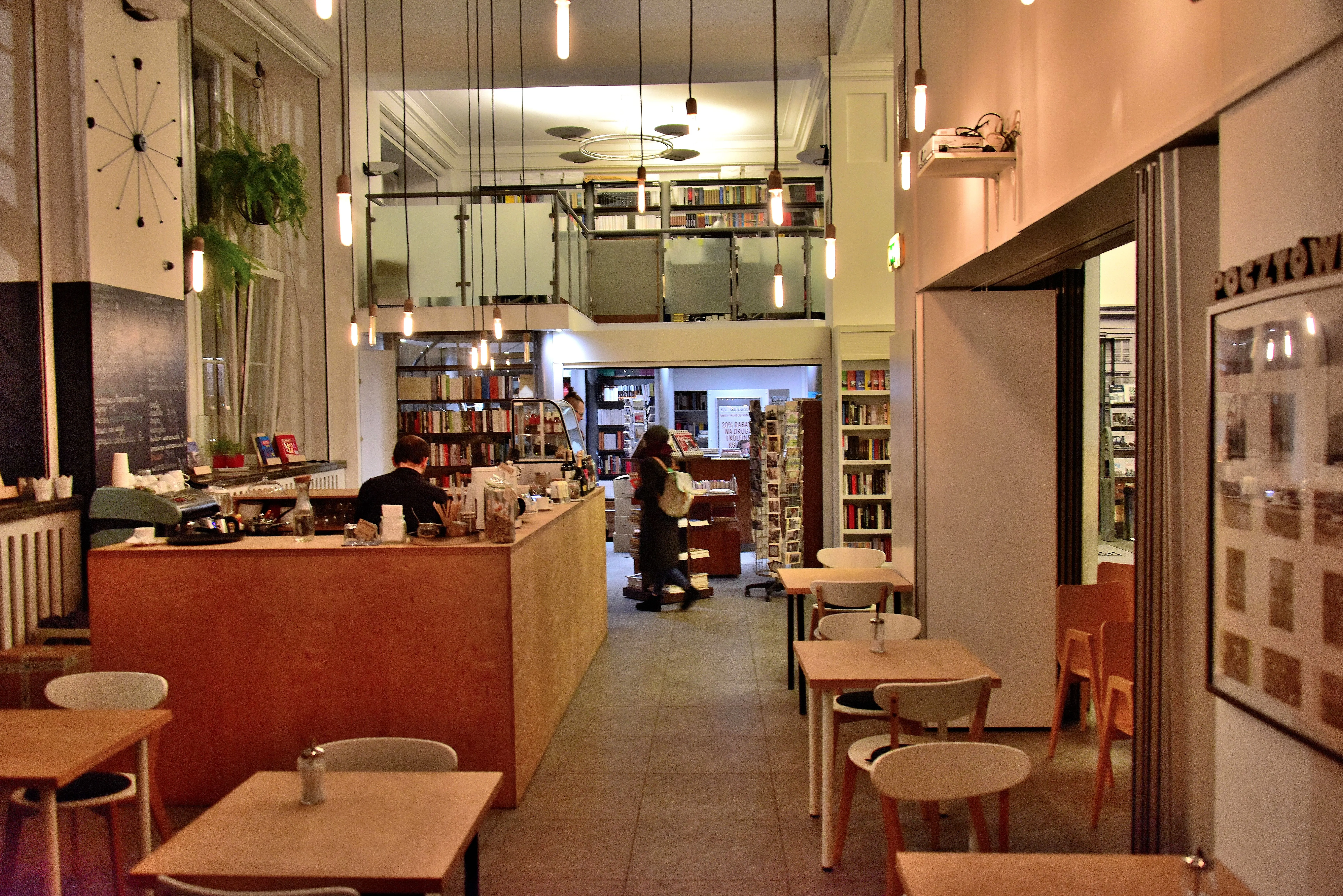 Interieur van een café met houten tafels, stoelen en boekenplanken, met een centrale toonbank en hangende lampen.