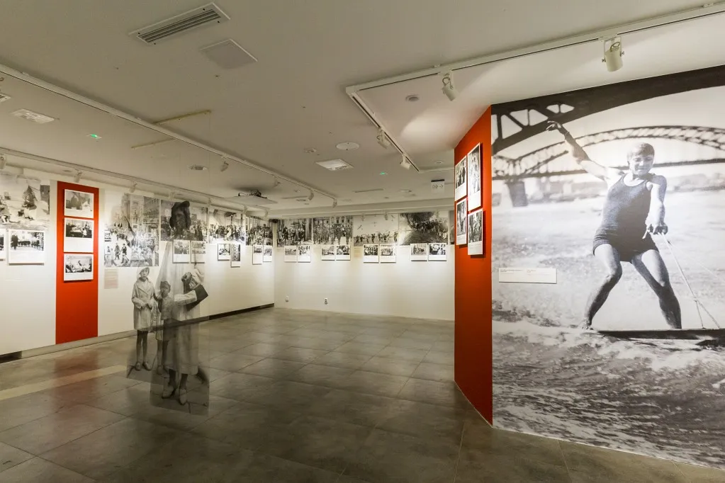 Interieur van een museumgalerij met witte muren, rode displaypanelen en grote zwart-witte historische foto's aan de muren.