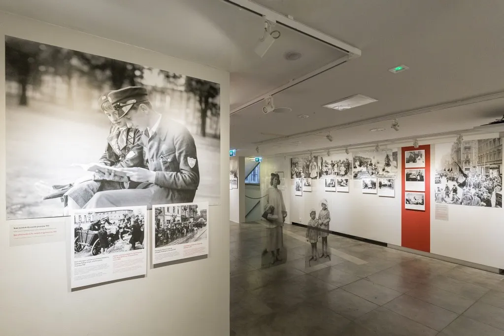 Tentoonstellingsgalerij met zwart-wit historische foto's aan de muren, waaronder een grote afbeelding van een soldaat die leest, en twee paspoppen op de achtergrond.