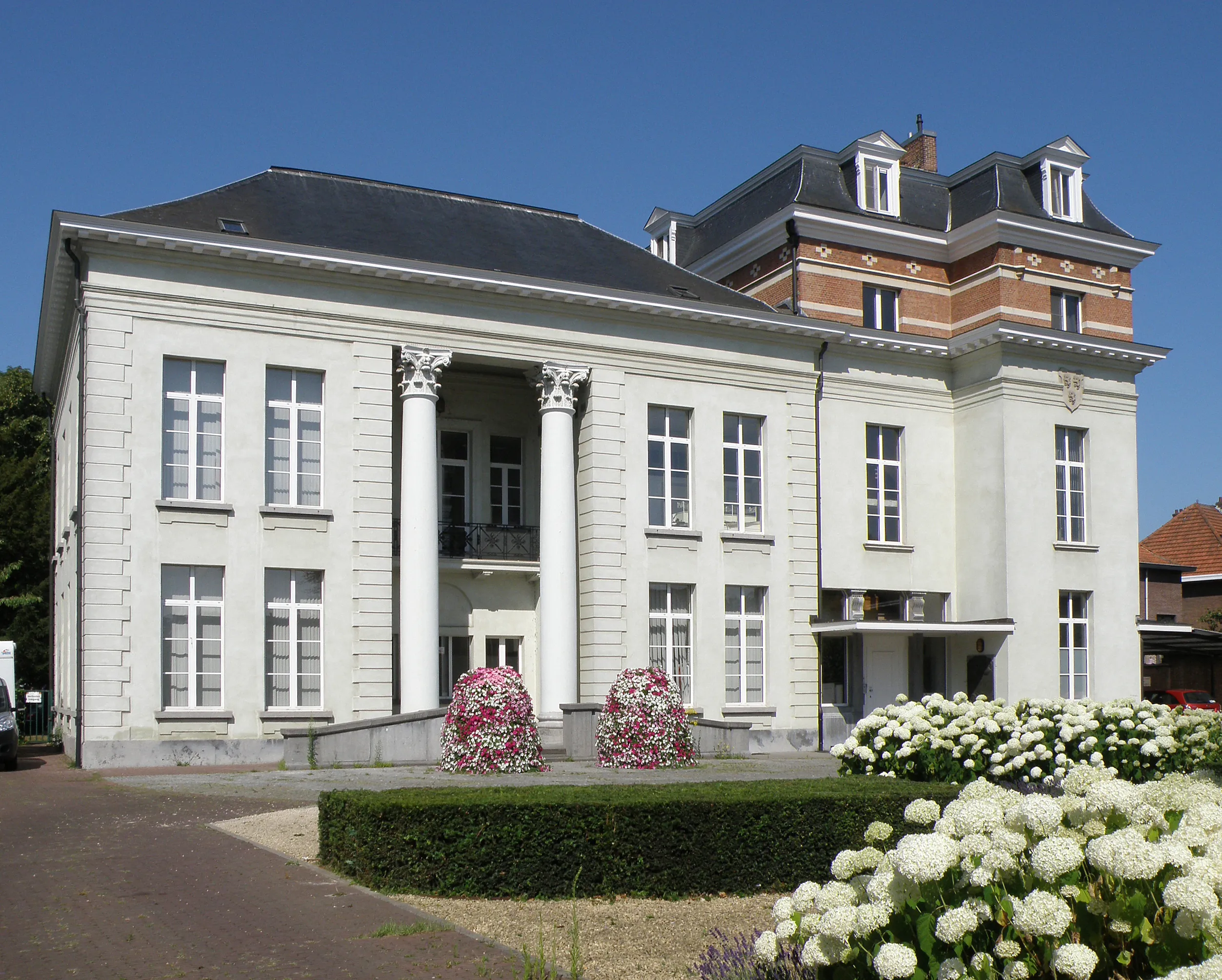 Vista exterior del Kasteel Broydenborg, un edificio clásico blanco con columnas en la entrada y una sección de torre de ladrillo con techo abuhardillado, con parterres en primer plano